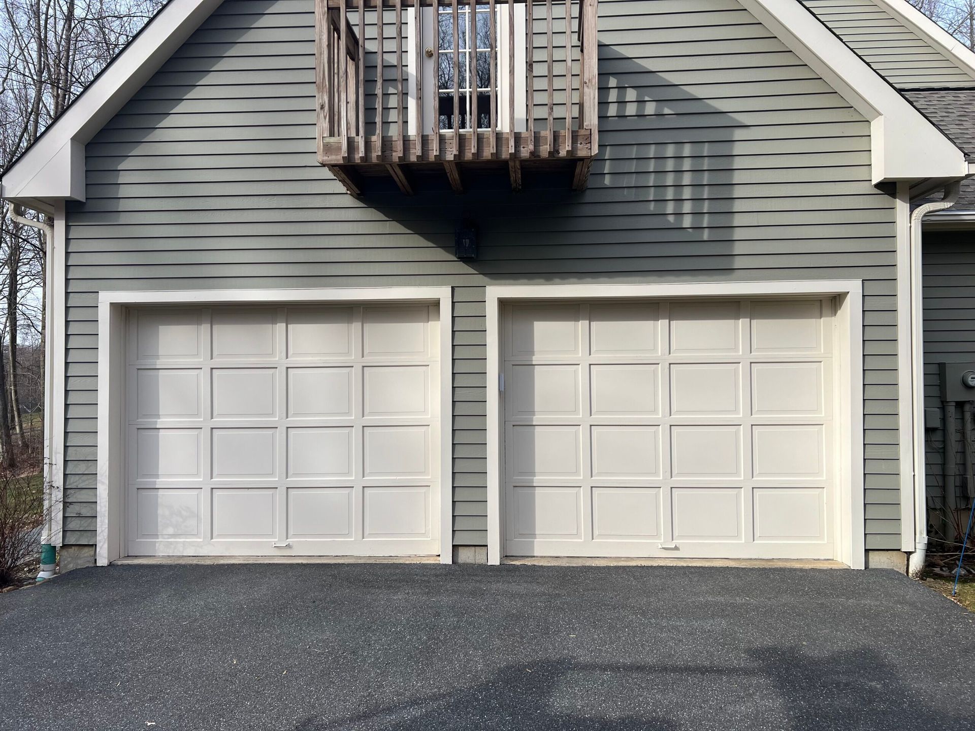 Two white garage doors on a gray house with a small balcony above and a black driveway