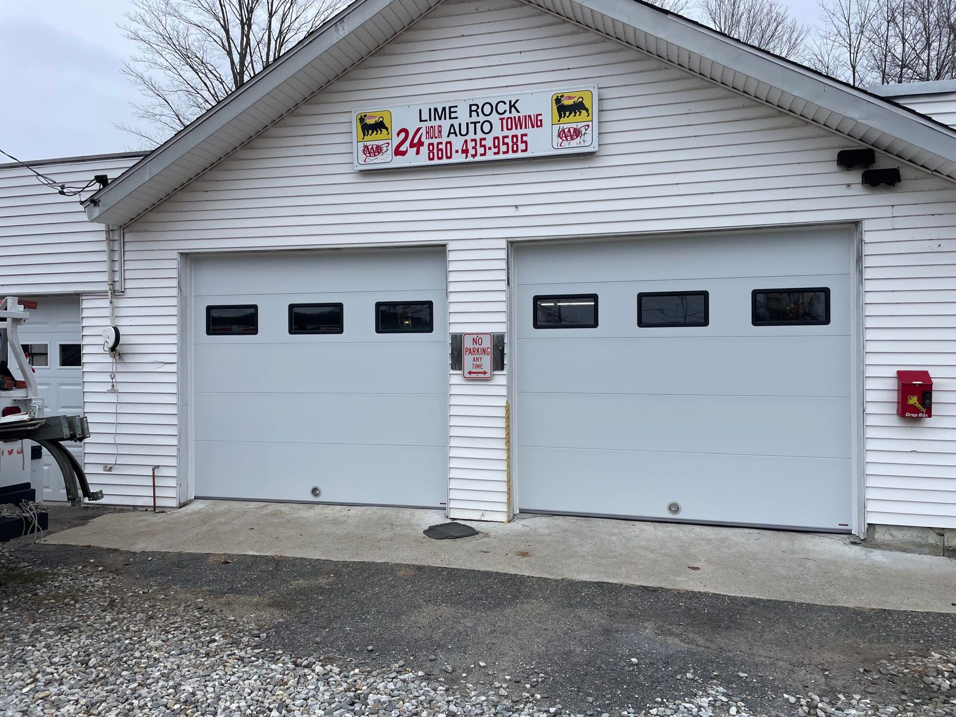 White two-car garage with closed doors and a sign above, on a gravel driveway beside a white house.
