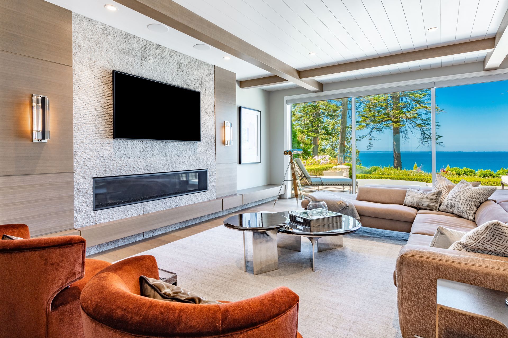 Living room with ocean view, modern design: fireplace, sectional sofa, orange chairs, large windows.