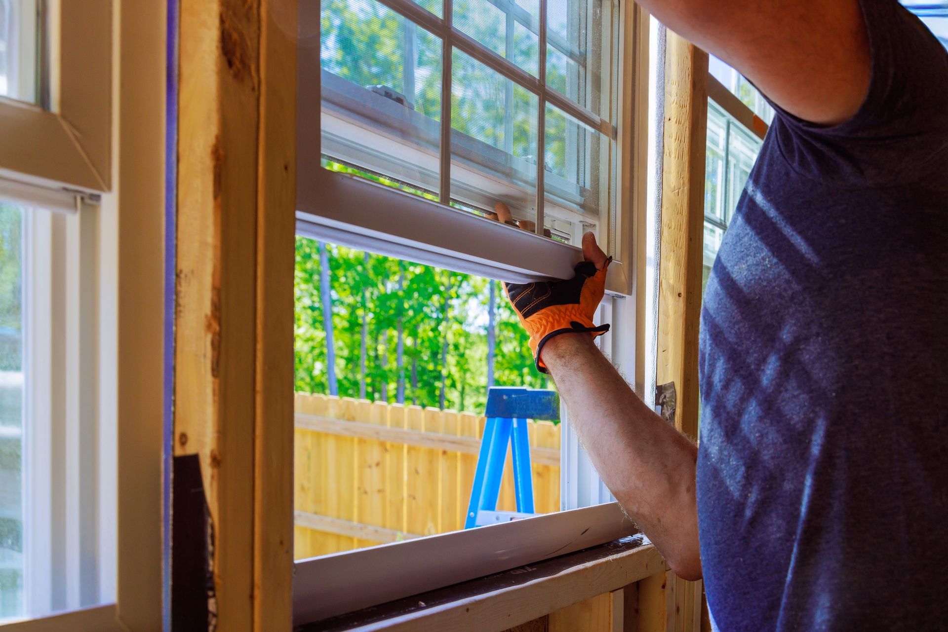 Man installing a window, wearing orange gloves. Sunlight streams through a green backdrop.
