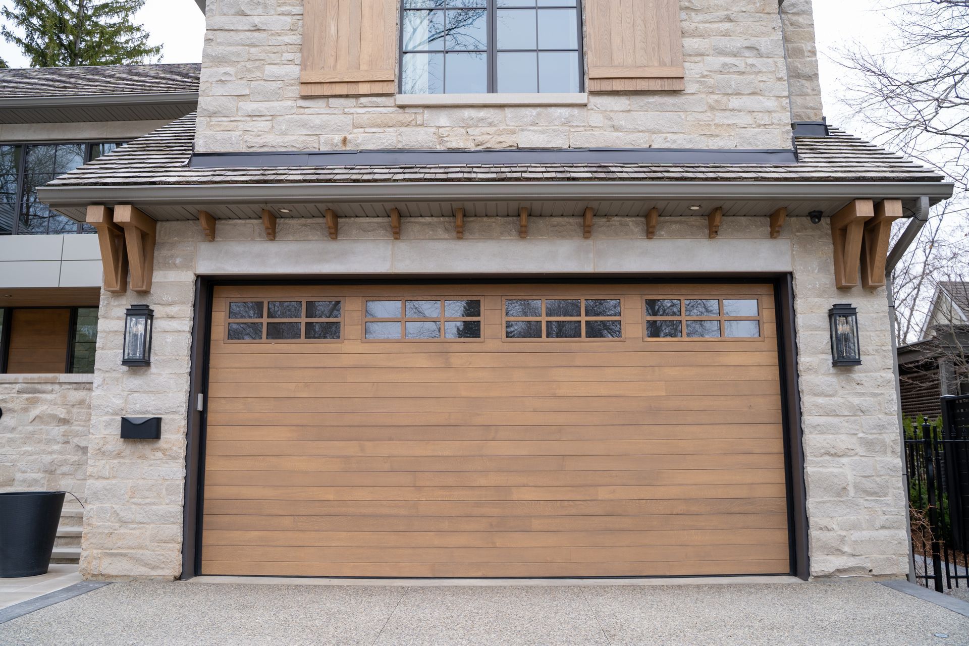 Tan wooden garage door with windows, flanked by stone walls and decorative lanterns.