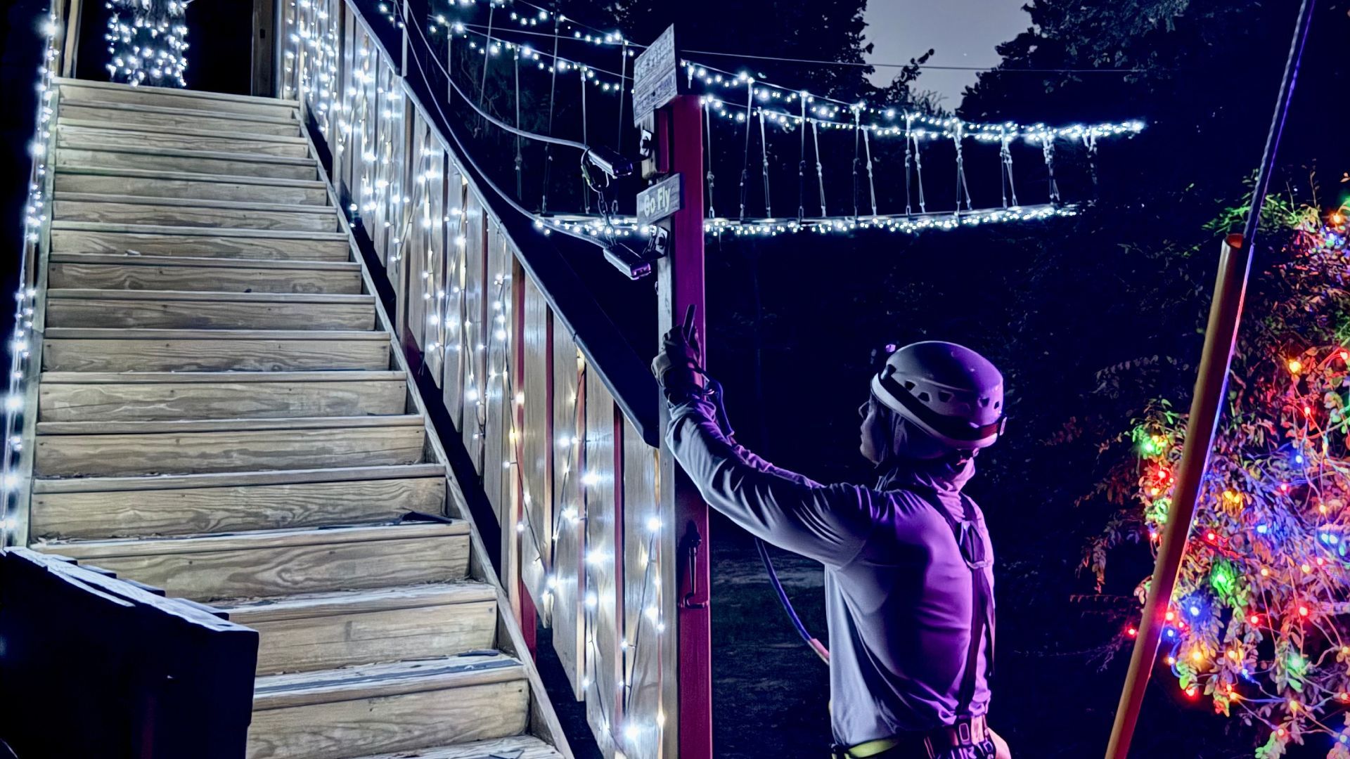 Person hanging holiday lights beside a wooden staircase at night