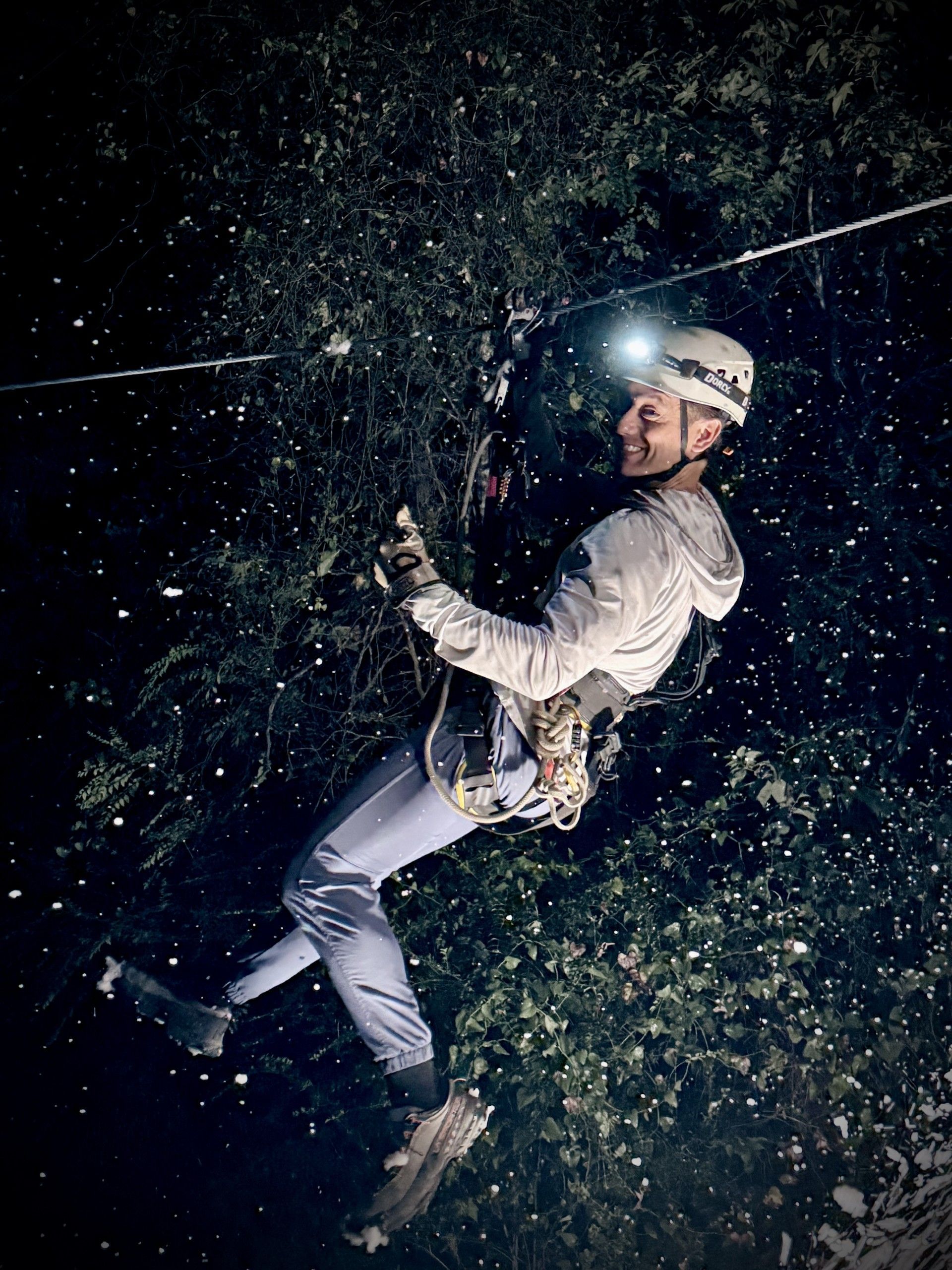 Person rappelling at night on a rope, wearing a helmet and white clothing, surrounded by falling water spray