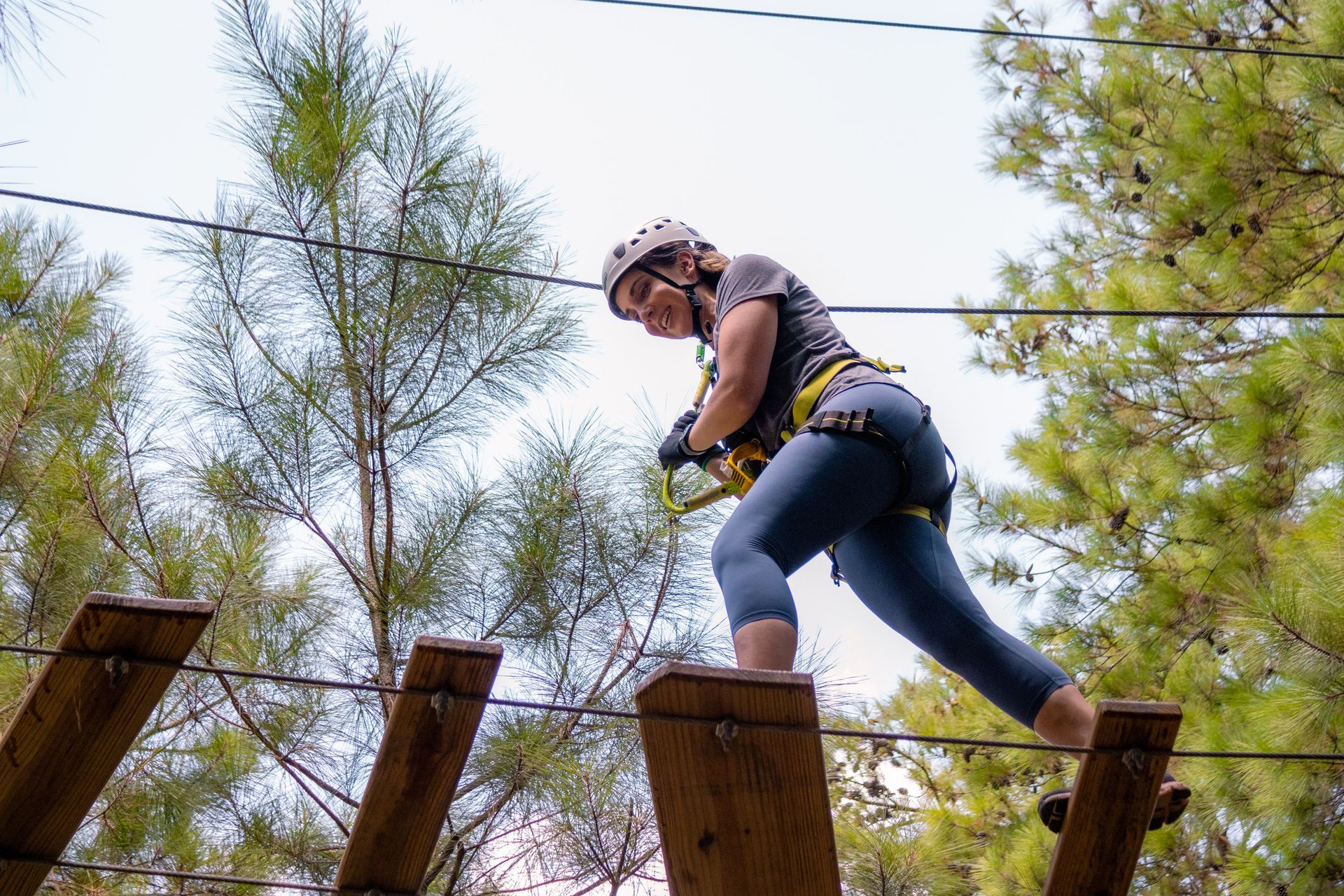 Person crossing a wooden ropes course outdoors, wearing a harness and helmet among trees.