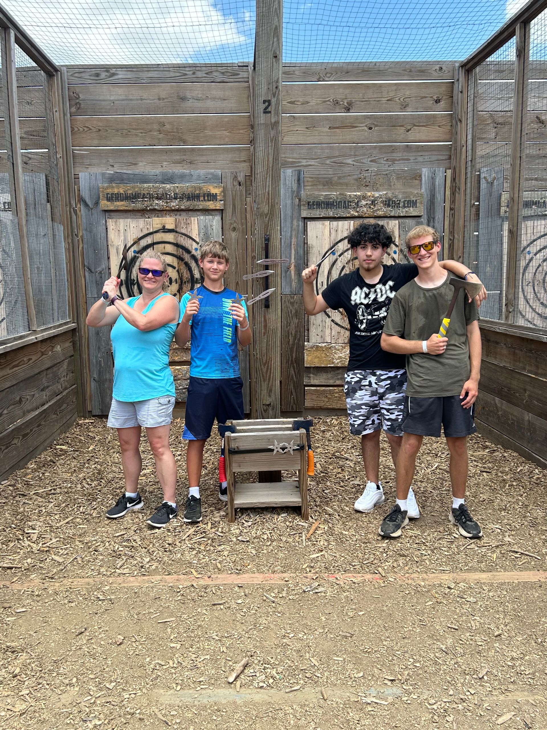 Four people posing and waving in front of a rustic wooden enclosure with a small crate on the ground.