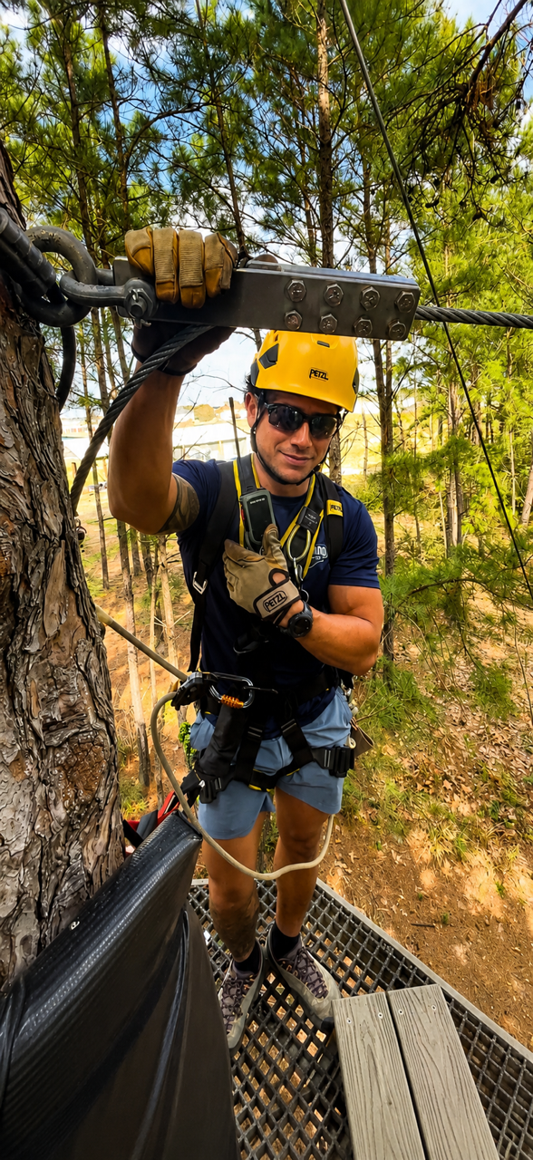 A climber in a yellow helmet and harness traverses a rope bridge in a forest canopy.
