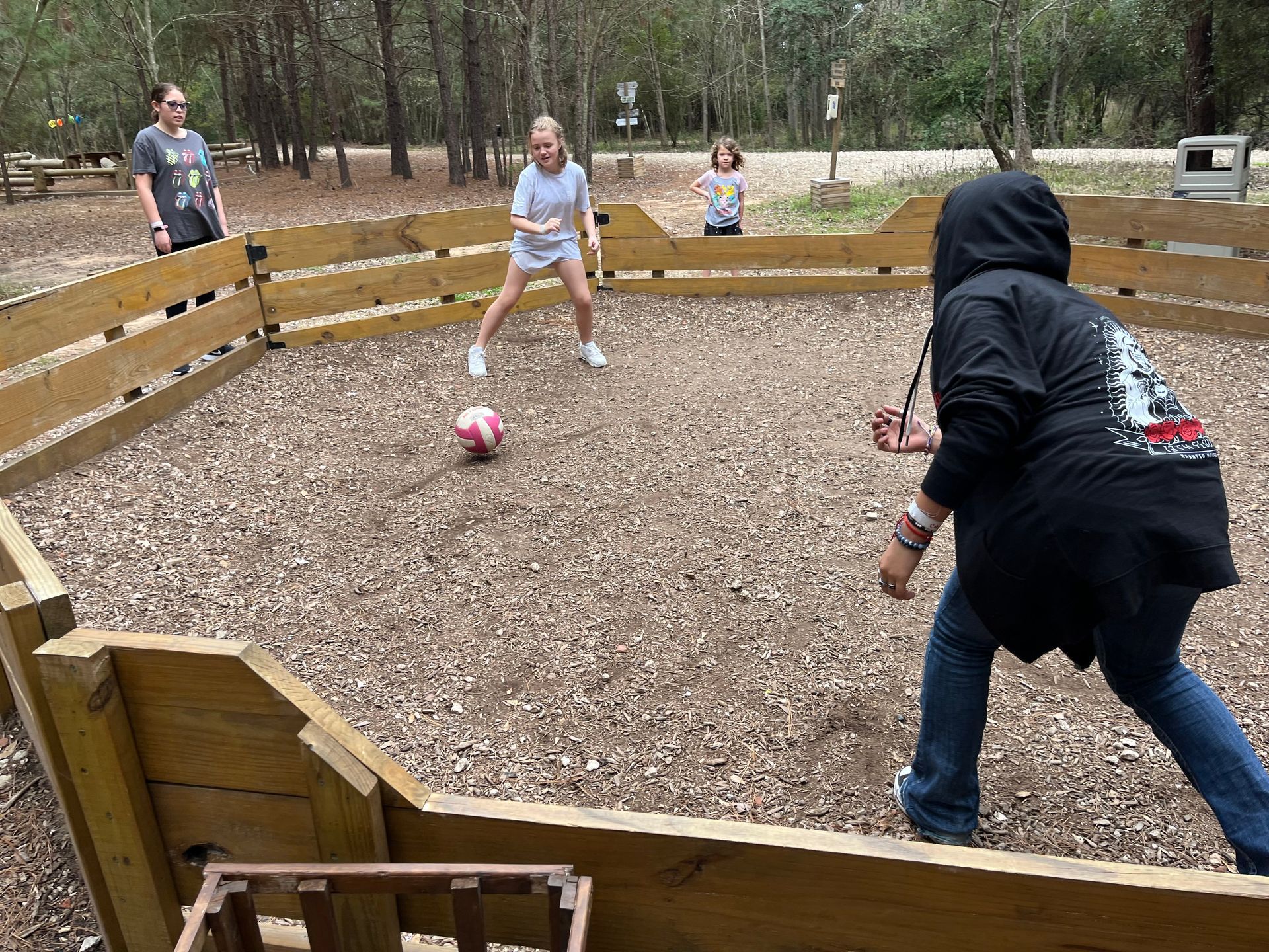 People playing a game of kickball in a wooded outdoor field, with a pink ball near center.