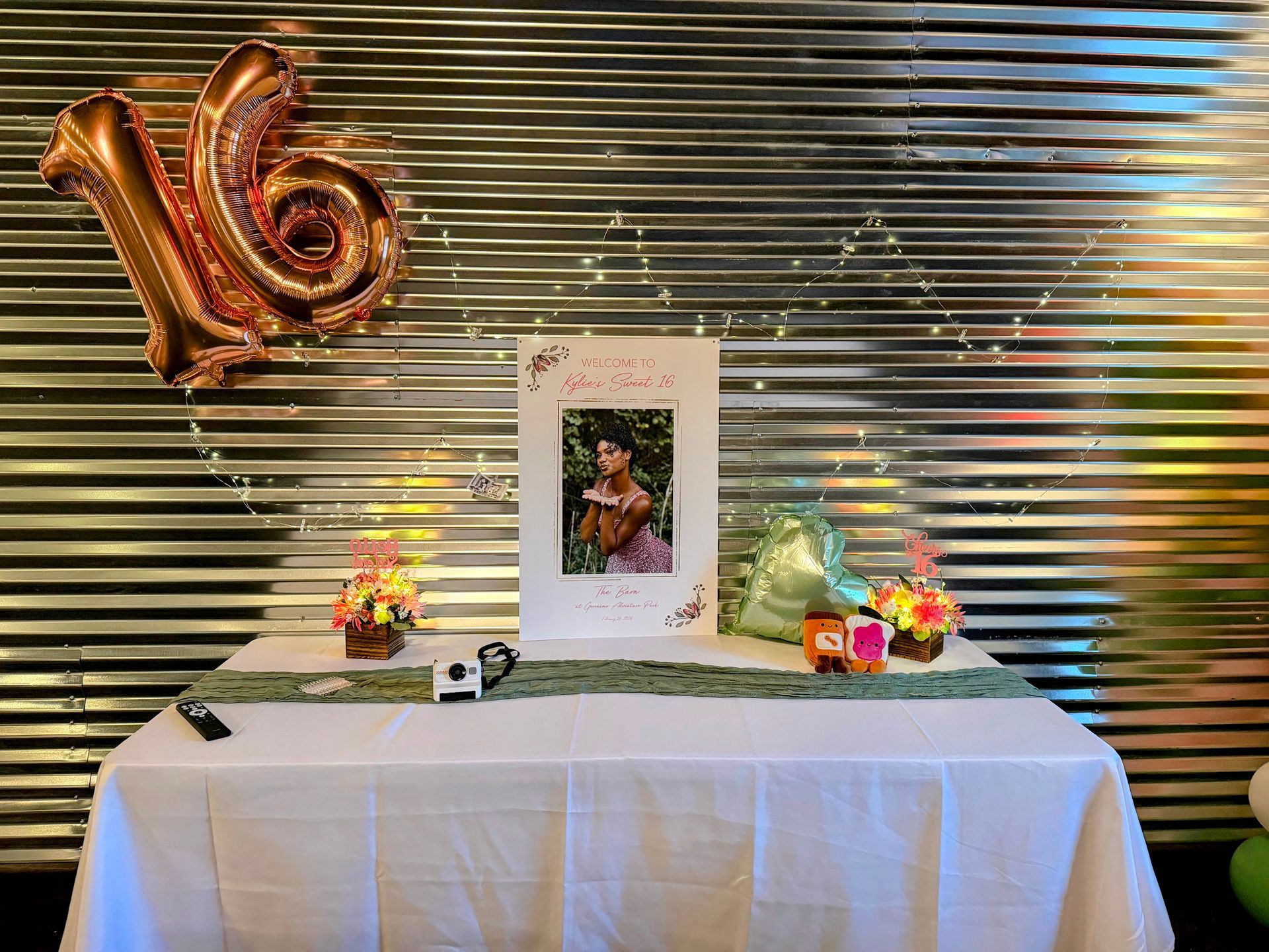 Decorated memorial table with framed portrait, flowers, candles, and gold “16” balloons against a silver backdrop
