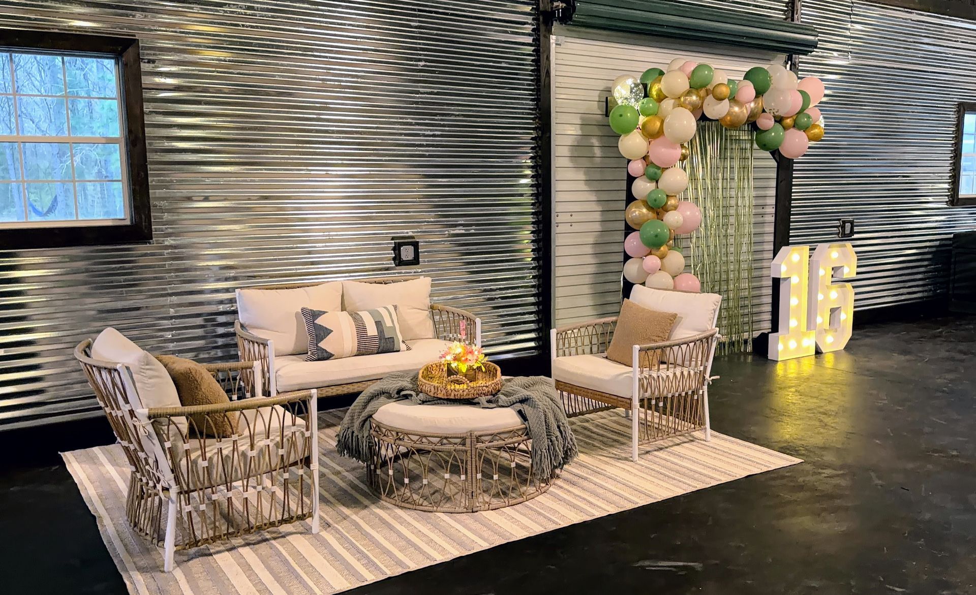 Cozy indoor lounge setup with wicker chairs, round coffee table, striped rug, and pastel balloon arch