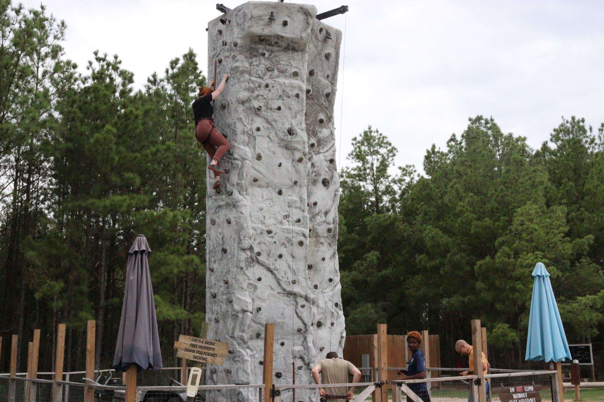 Person climbing a tall outdoor rock wall at a recreational park, with trees and umbrellas nearby