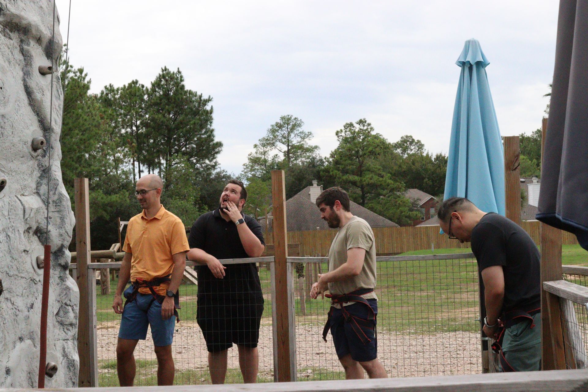 Four men chatting beside a pool, with a rock wall and a closed blue umbrella in the background.