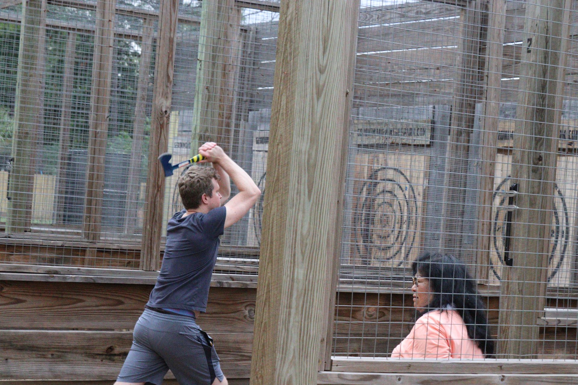 Person aiming a bow in a netted outdoor target area while another sits nearby.