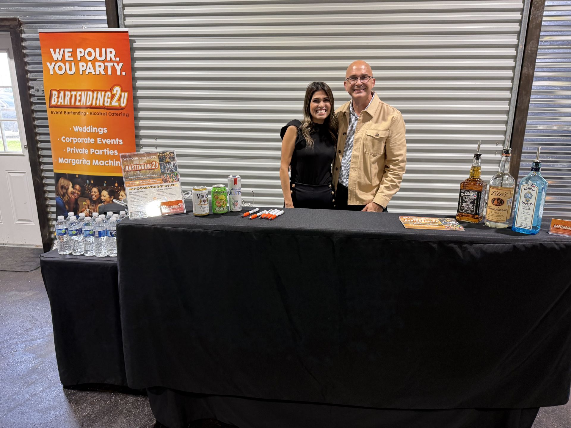 Two people standing behind a black-draped table at a vendor booth with product displays and a promotional sign.