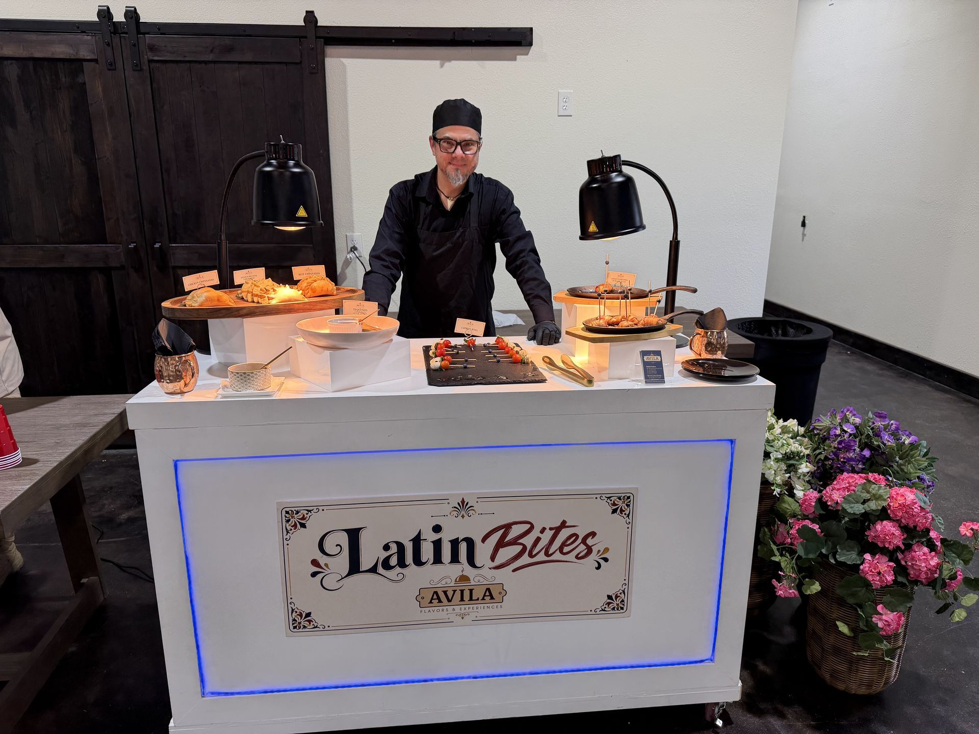 Buffet vendor behind LatinBites table with warm food, lamps, flowers, and pink bouquets on the side.