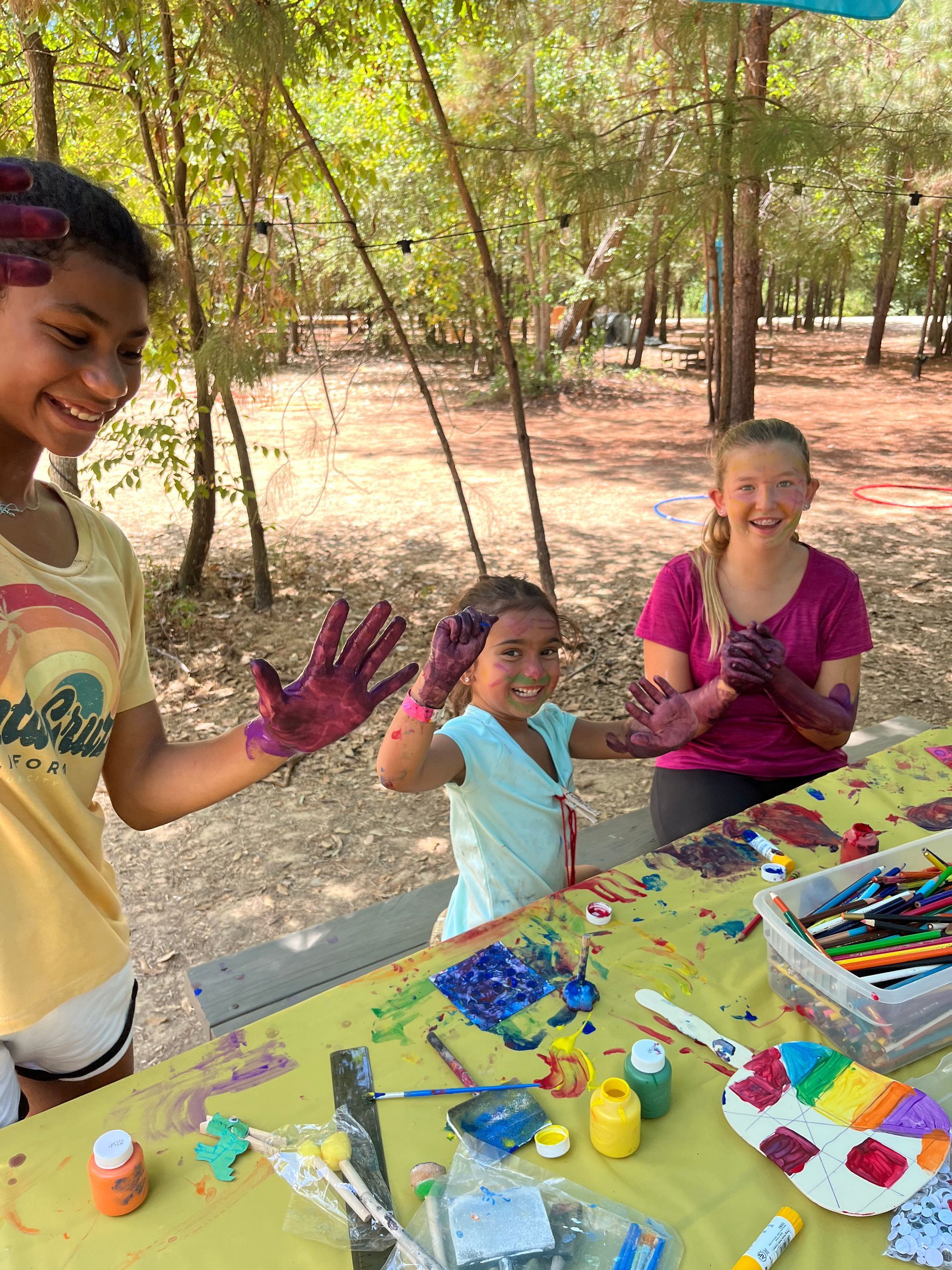 Children painting at an outdoor art table, with colorful supplies and paint-covered hands under trees.