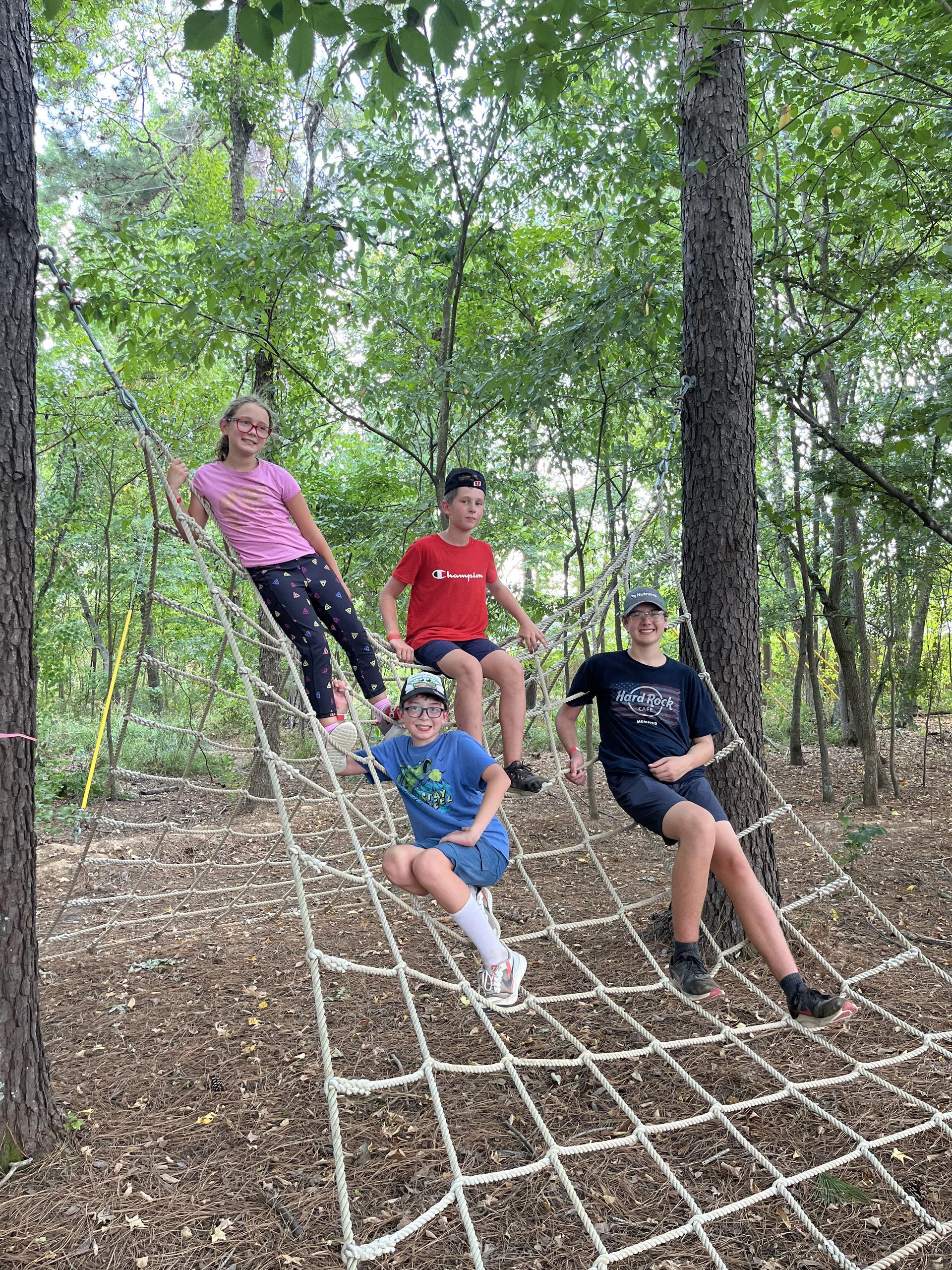 Four children posing on a rope net in a wooded area, wearing colorful clothes.