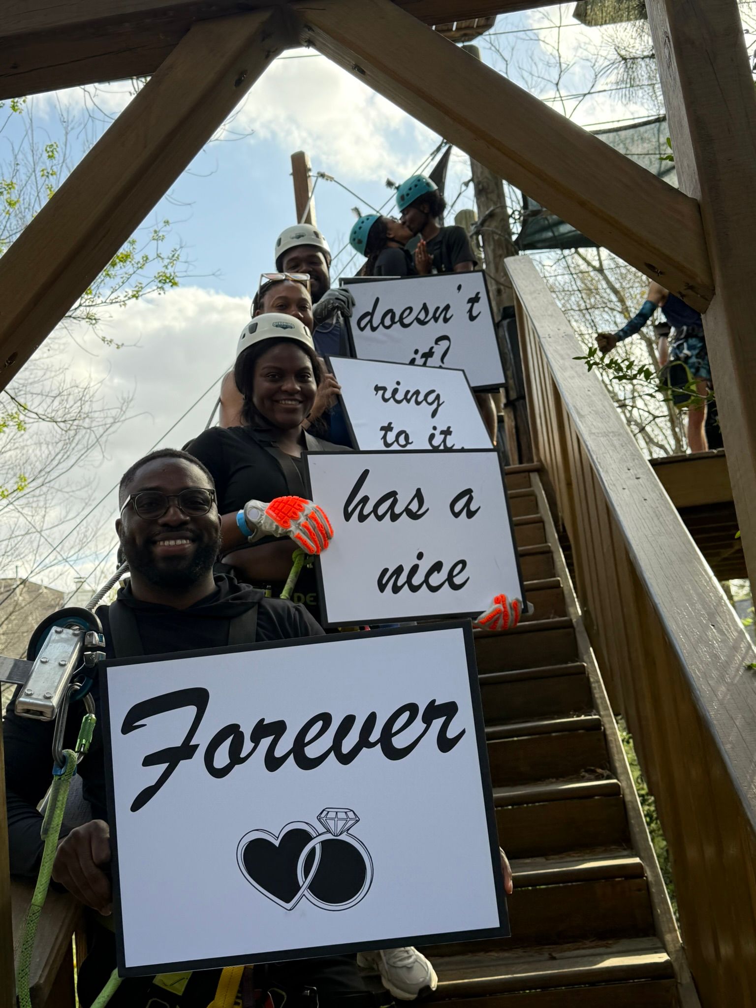 Three people on stairs hold signs reading “Forever,” “has a nice,” and “doesn’t have a nice” in a wooden structure.