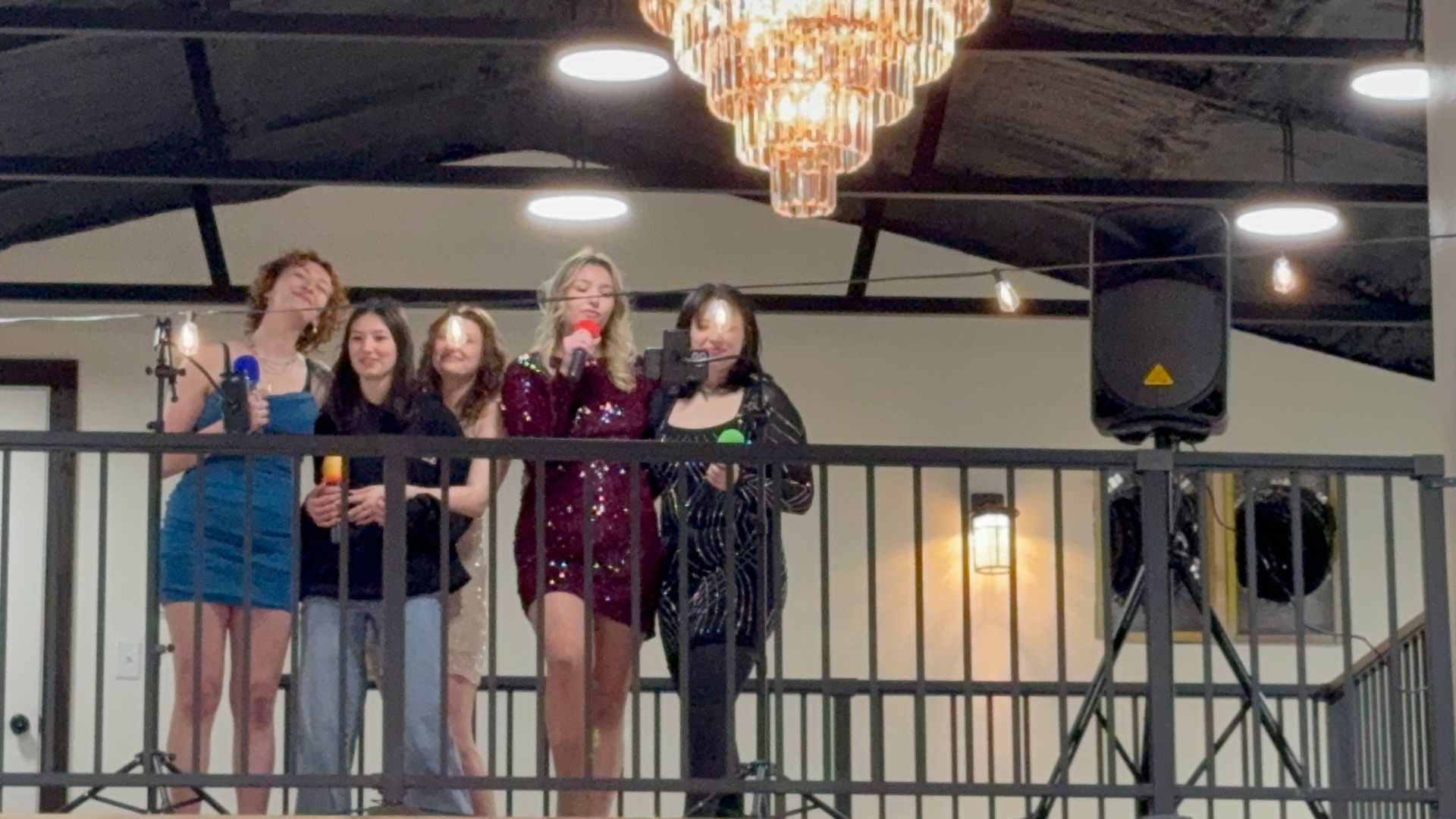 Four people stand on a balcony, singing into microphones under a chandelier.