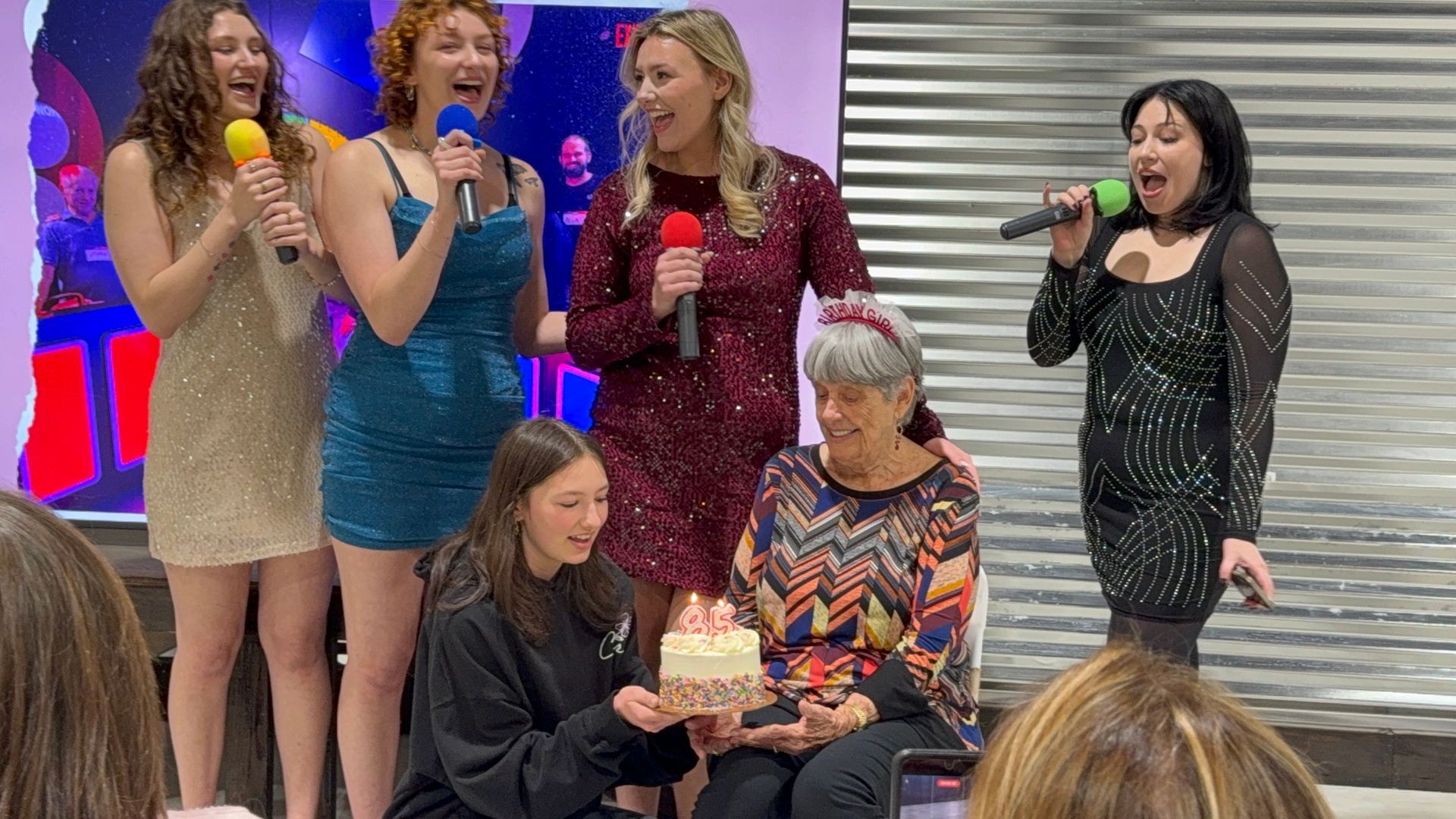 Five women onstage singing into colorful microphones, with two kneeling in front and a bright backdrop.