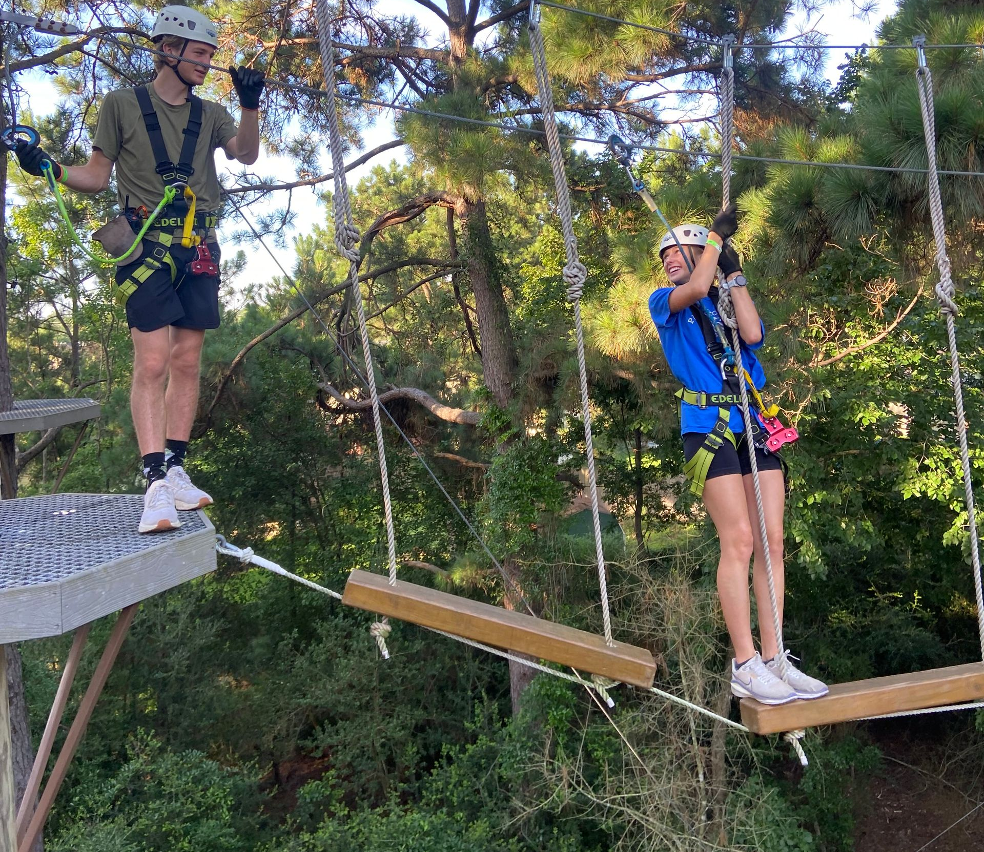 Two people wearing harnesses cross a rope bridge in a forest adventure course