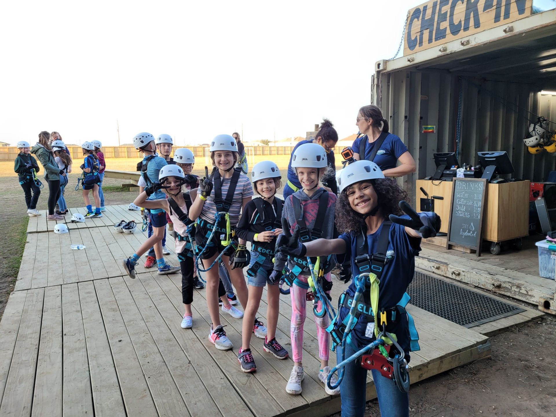 Children in helmets and harnesses line up at an outdoor zipline course, holding ropes near a check-in area