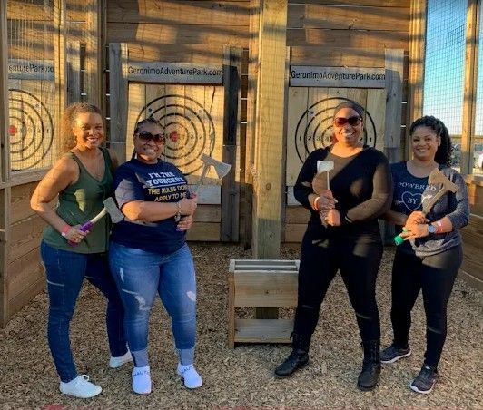Four people pose outdoors at an axe-throwing range, each holding a drink and smiling.