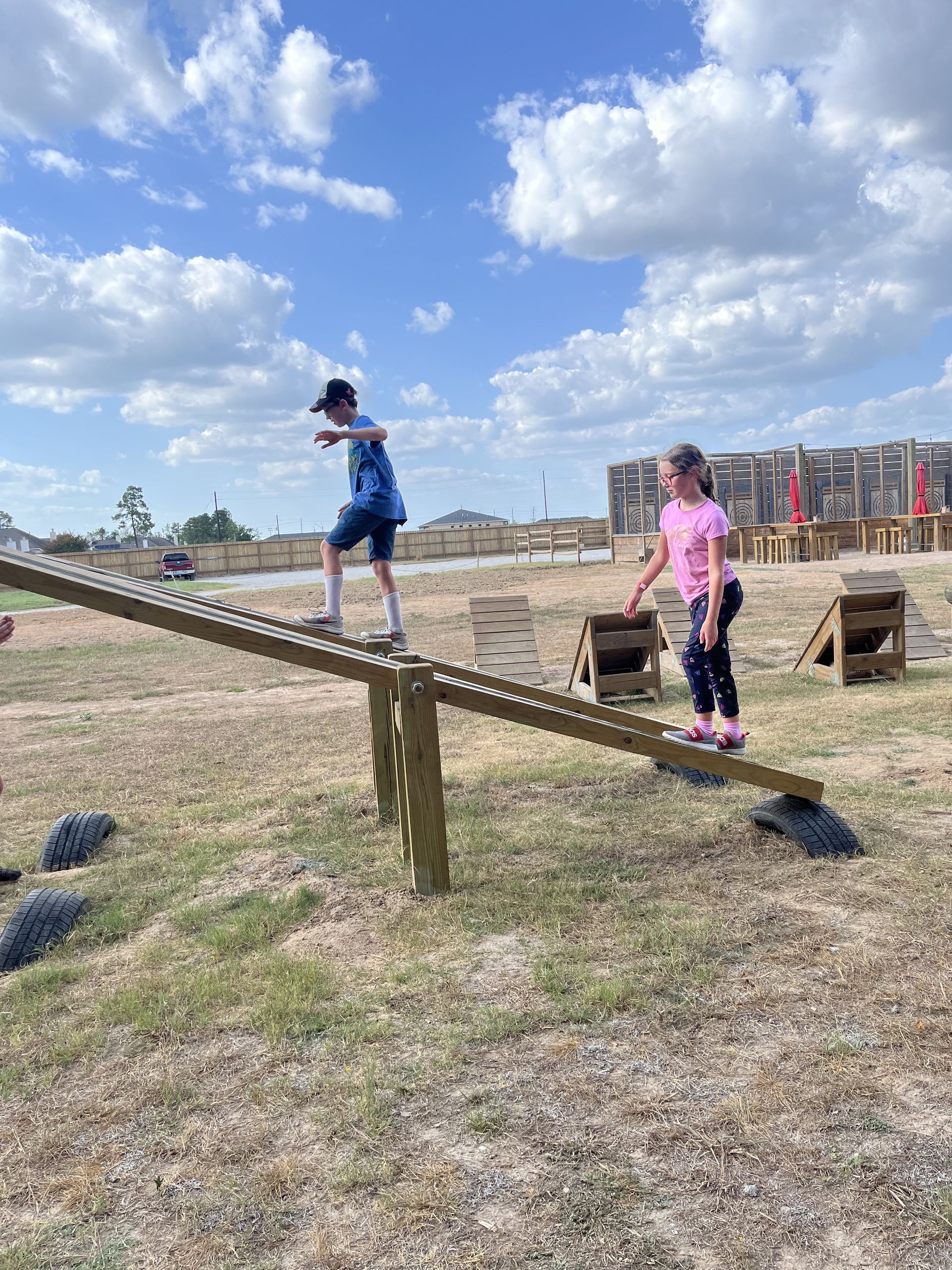 Two children balancing on a slanted wooden beam in a grassy park under a partly cloudy sky