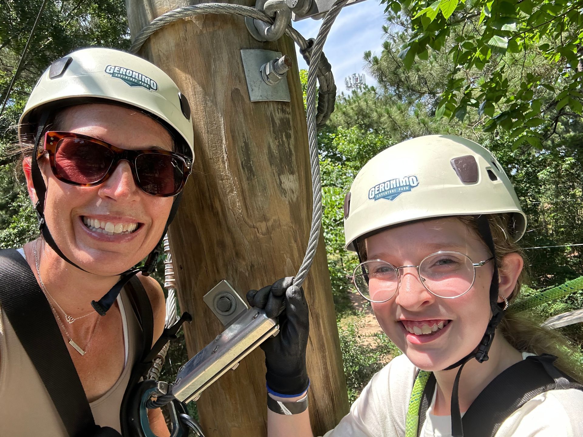 Two climbers smiling beside a tree trunk, wearing helmets, harnesses, and gloves outdoors.