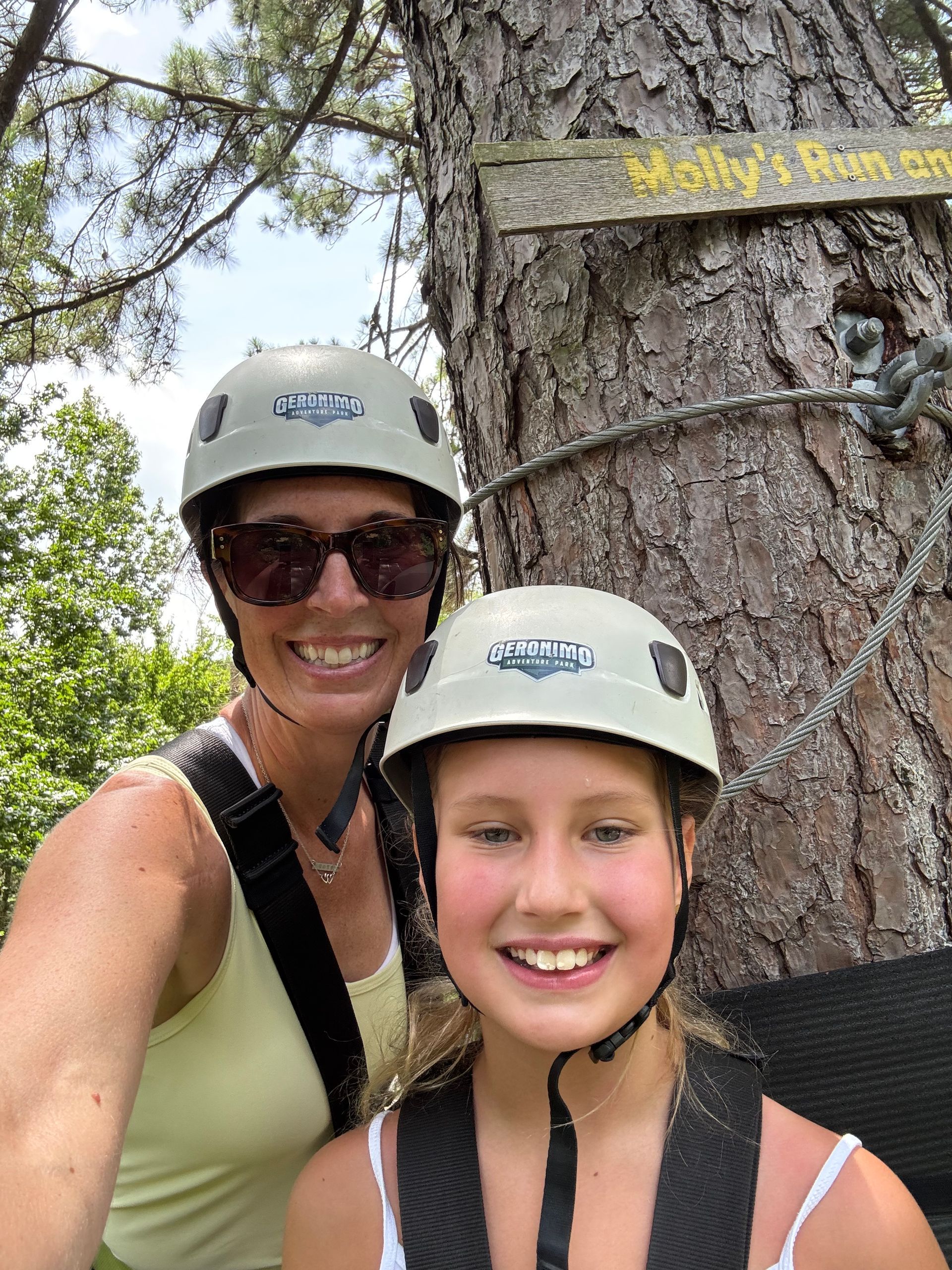 Two people smiling in helmets and life vests outdoors by a tree, likely on a zipline adventure.