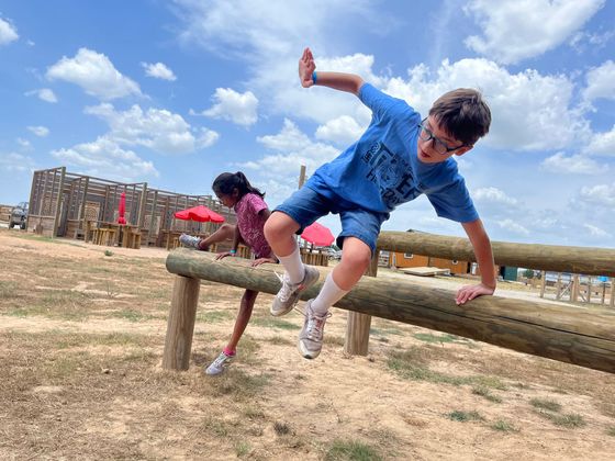 Two children climbing over a wooden obstacle beam in a sandy outdoor play area under a blue sky
