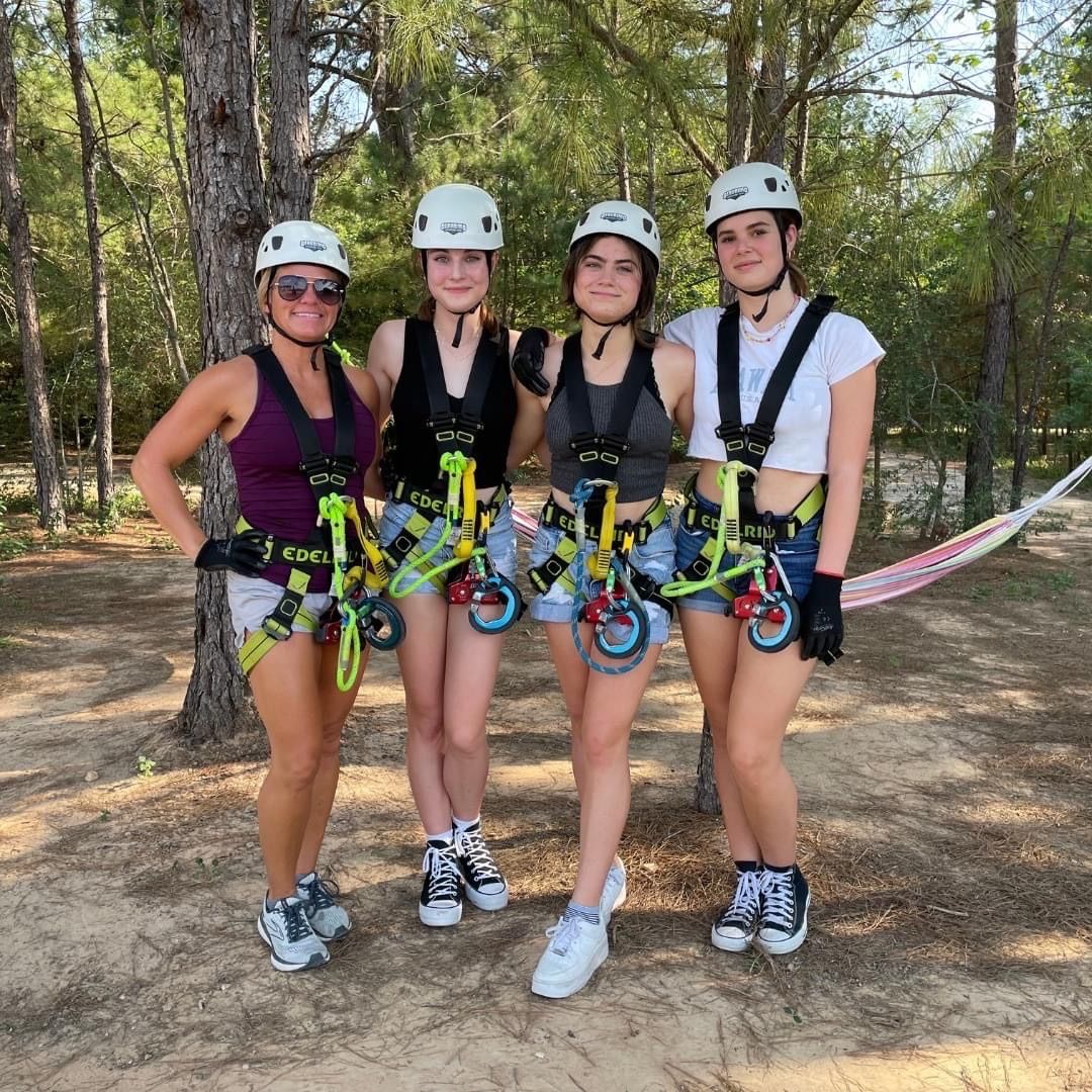 Four people in climbing gear and helmets posing outdoors on a wooded trail.