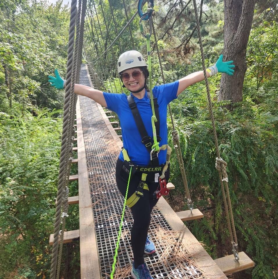 Person in helmet and harness smiling on a suspension bridge in a lush forest, arms outstretched