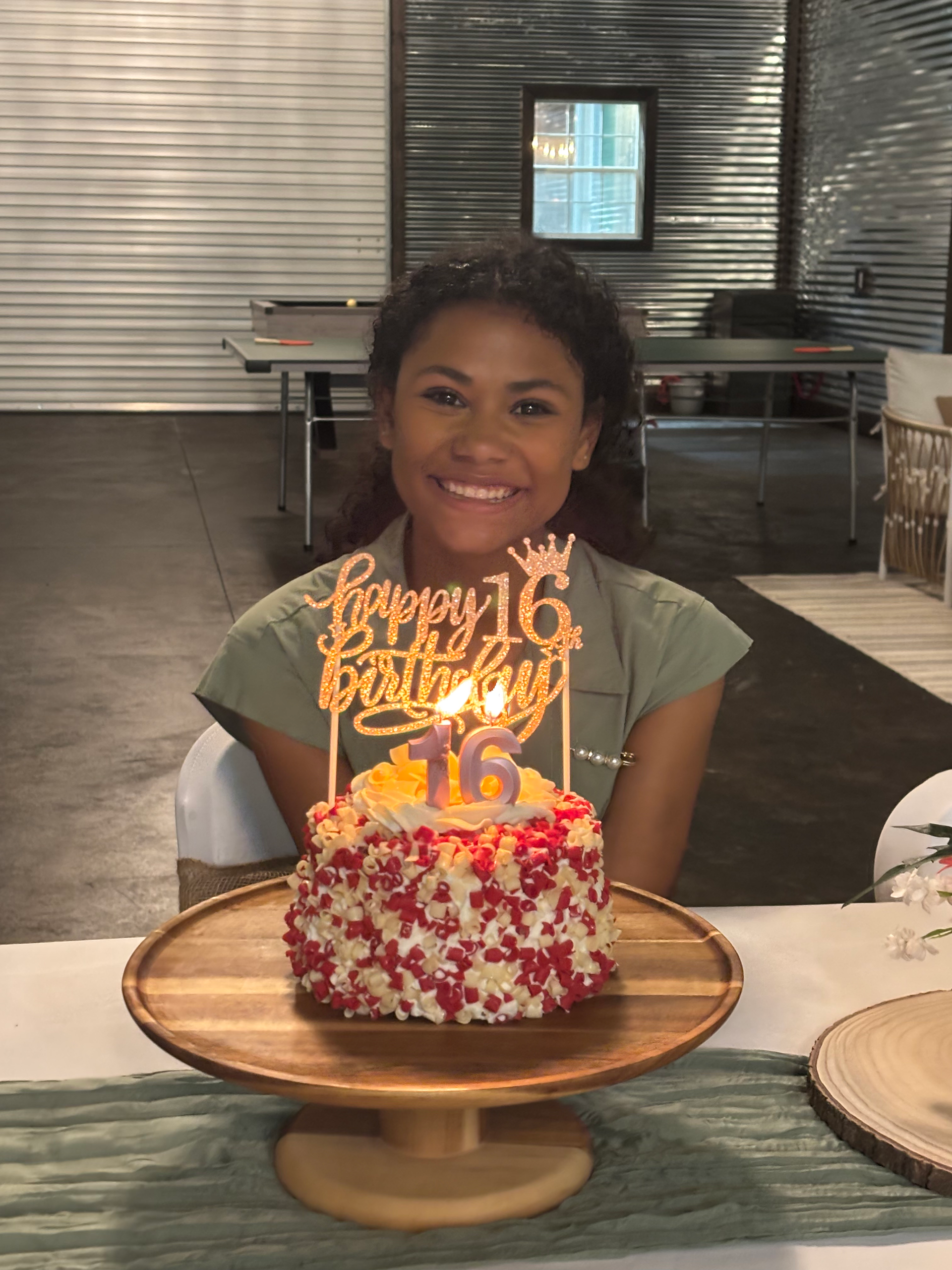 Person smiling behind a birthday cake with lit candles and a “Happy Birthday” topper in a restaurant.