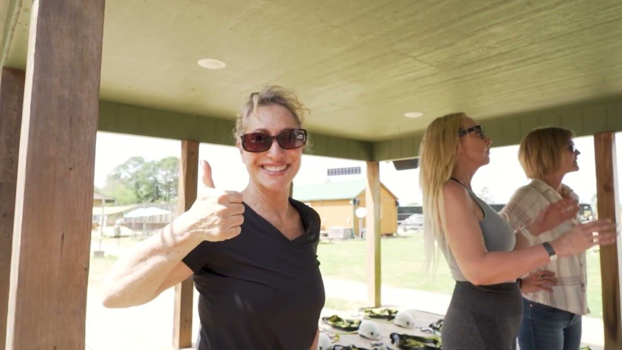 Woman in sunglasses smiling and giving a thumbs-up at an outdoor pavilion, with two people in the background.