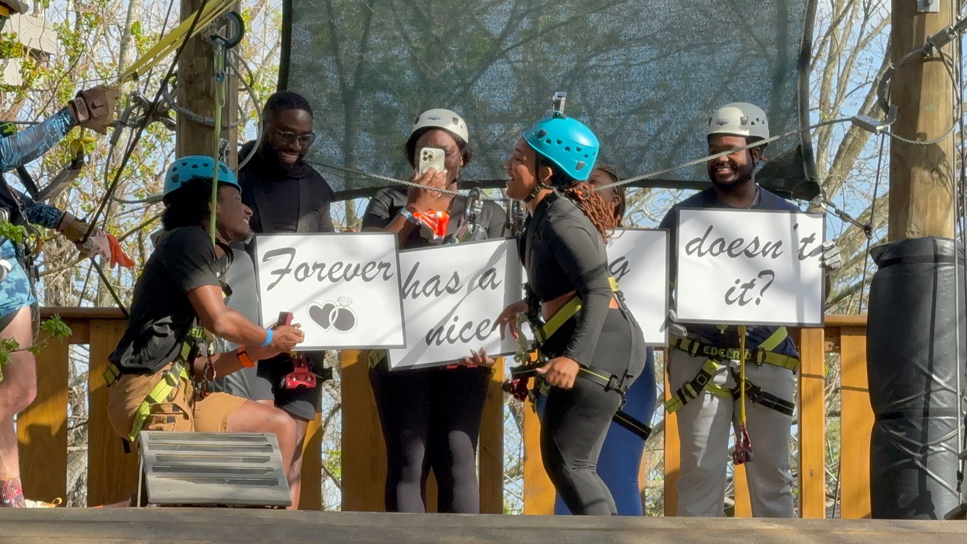 Five people stand outdoors holding protest signs and wearing helmets near a tree-lined area.