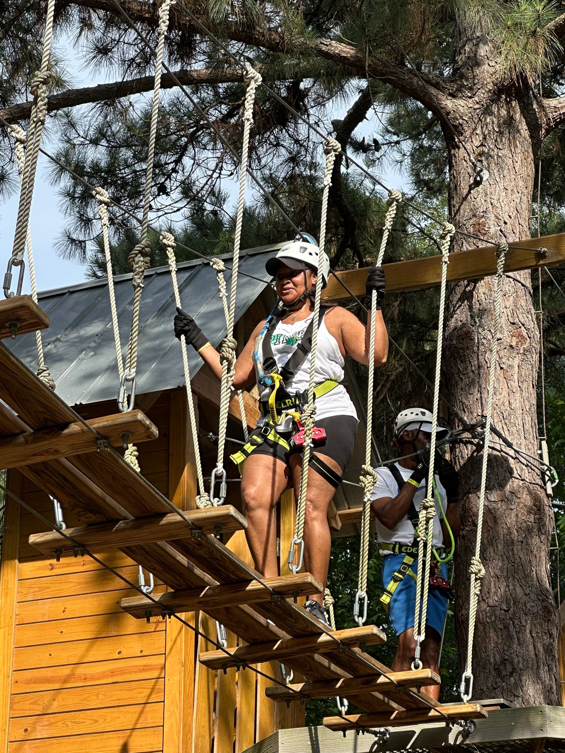 Two people crossing a rope bridge at an outdoor adventure park, wearing harnesses and helmets.