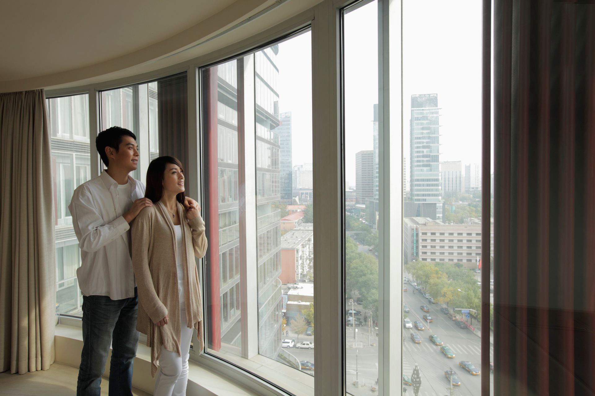 Couple gazing out a curved window at a city street and buildings. The man has his arm around the woman.
