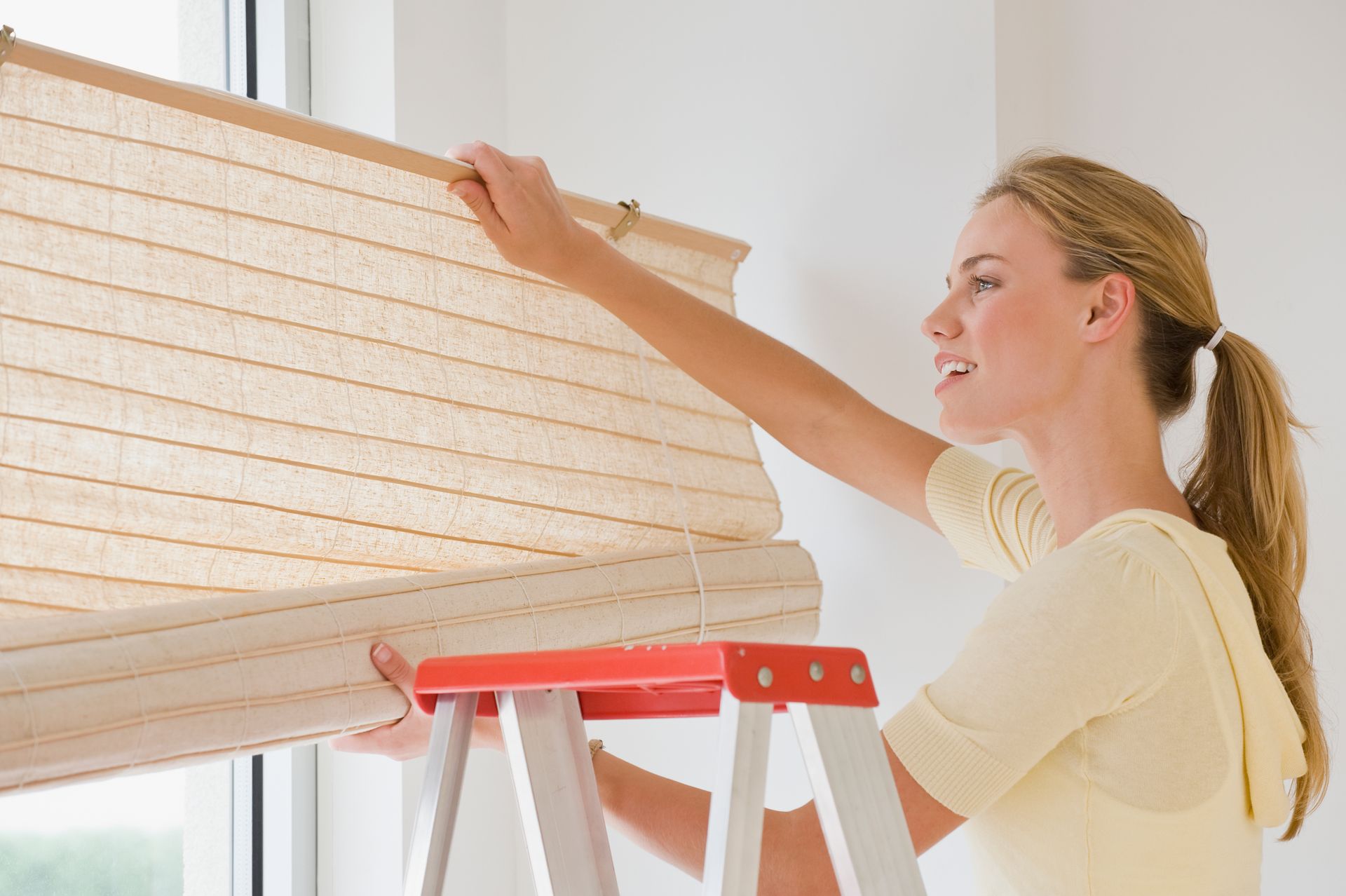 Blonde woman on a ladder adjusting beige roman shade by white wall, Custom Window Treatments by Blin