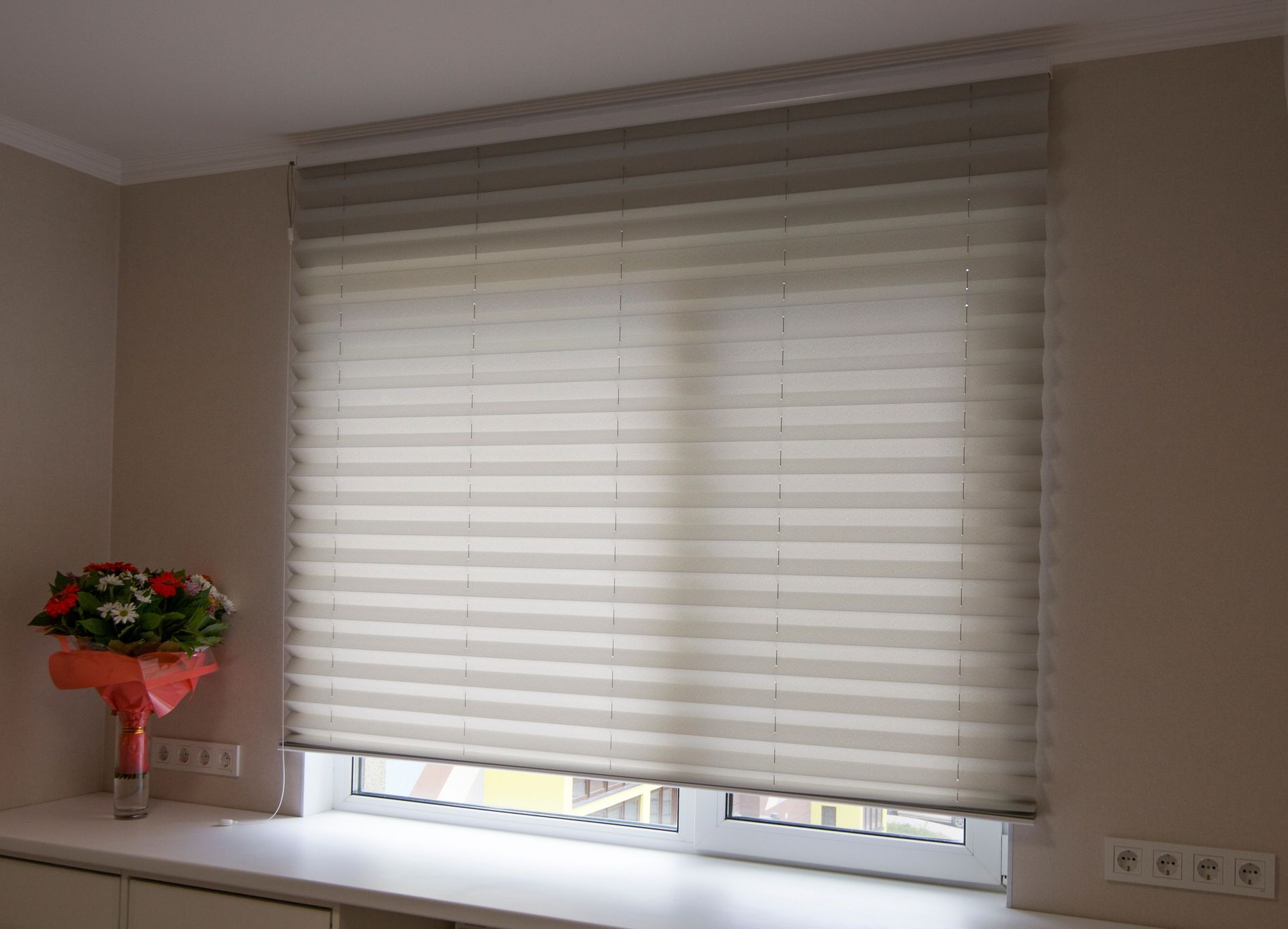 Closed layered window blinds above a sill with flowers and power outlets inside a room. Closed layered window blinds above a sill with flowers and power outlets inside a room.