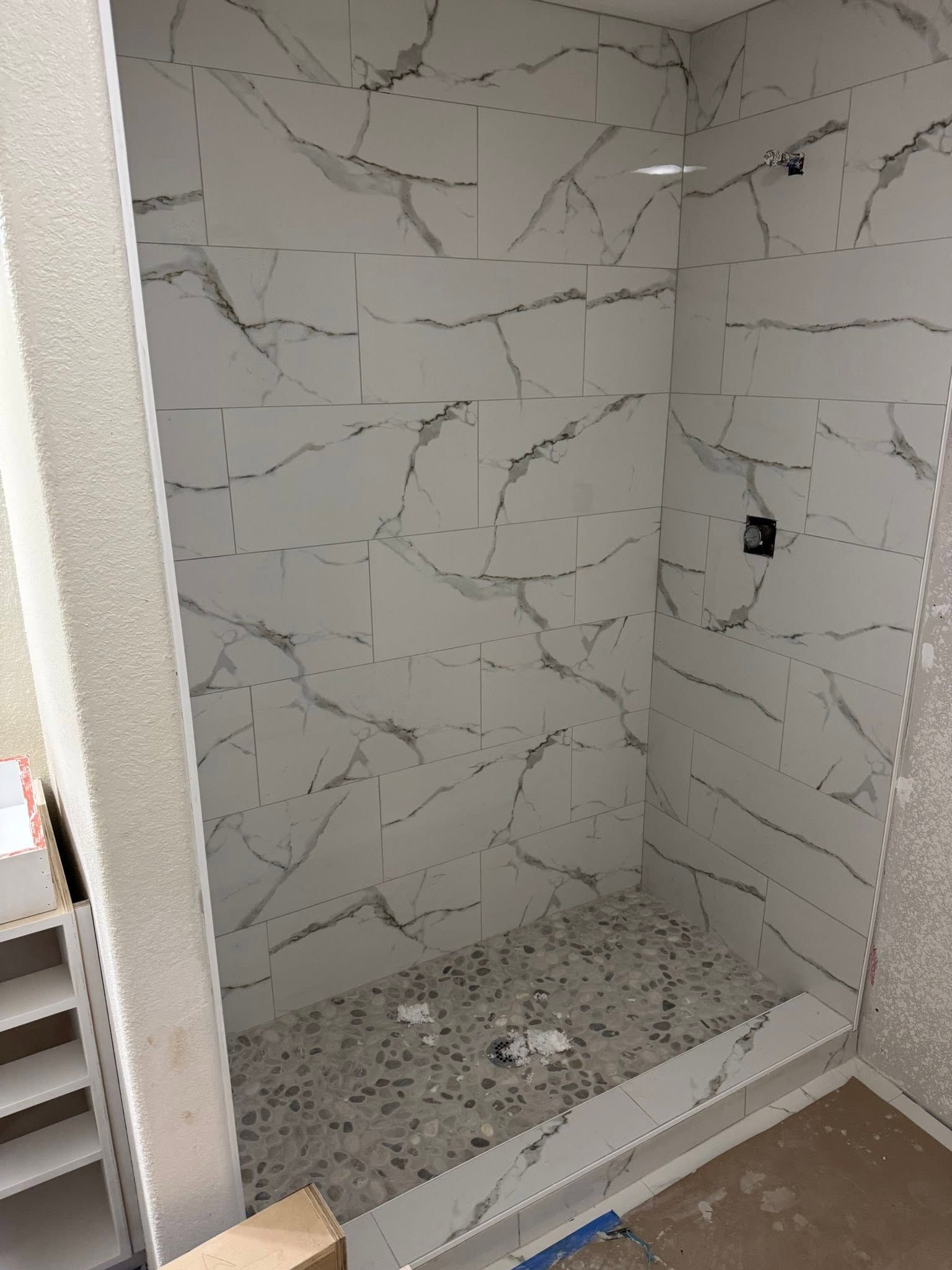 Partially tiled shower alcove with marble-look walls, pebble floor, and a low curb under construction