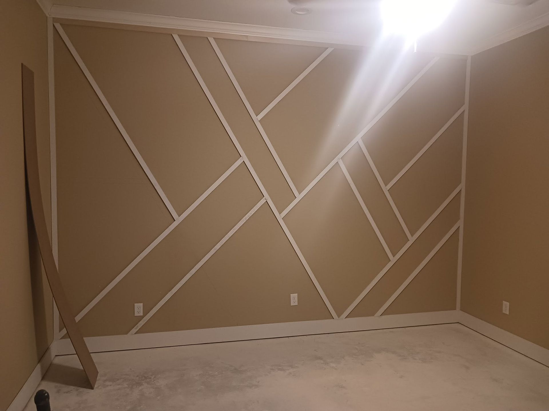 Empty beige room with white diagonal tape lines on one wall and a floor tarp under a bright ceiling light