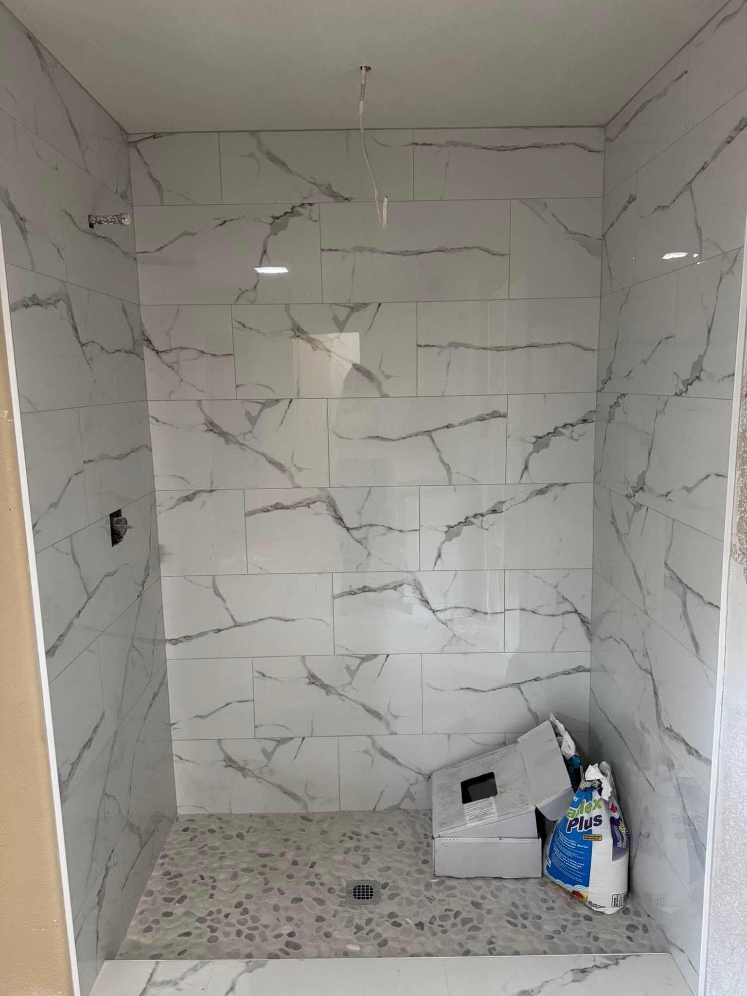 Empty tiled shower stall with white marble-look walls and a speckled floor, with cleaning supplies in the corner