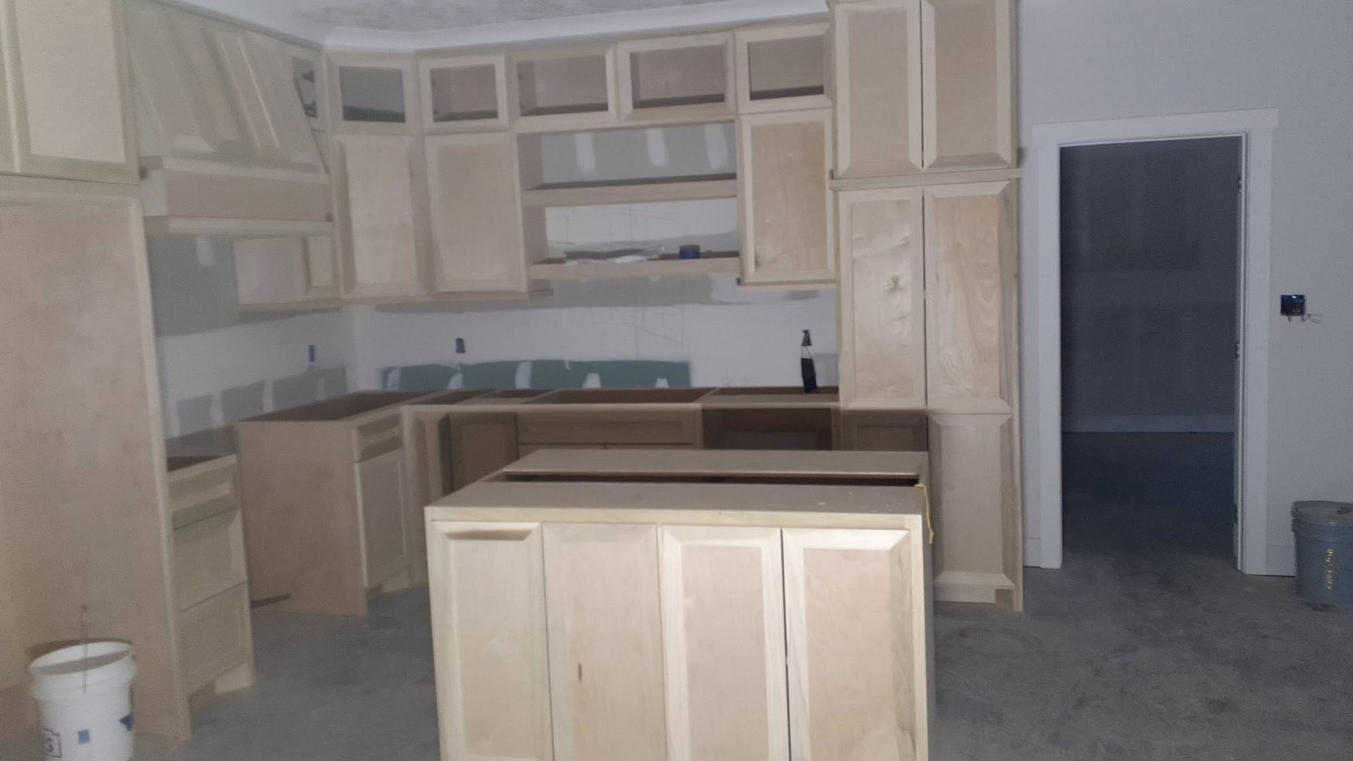 Unfinished kitchen with light wood cabinets, island, and gray flooring under construction.