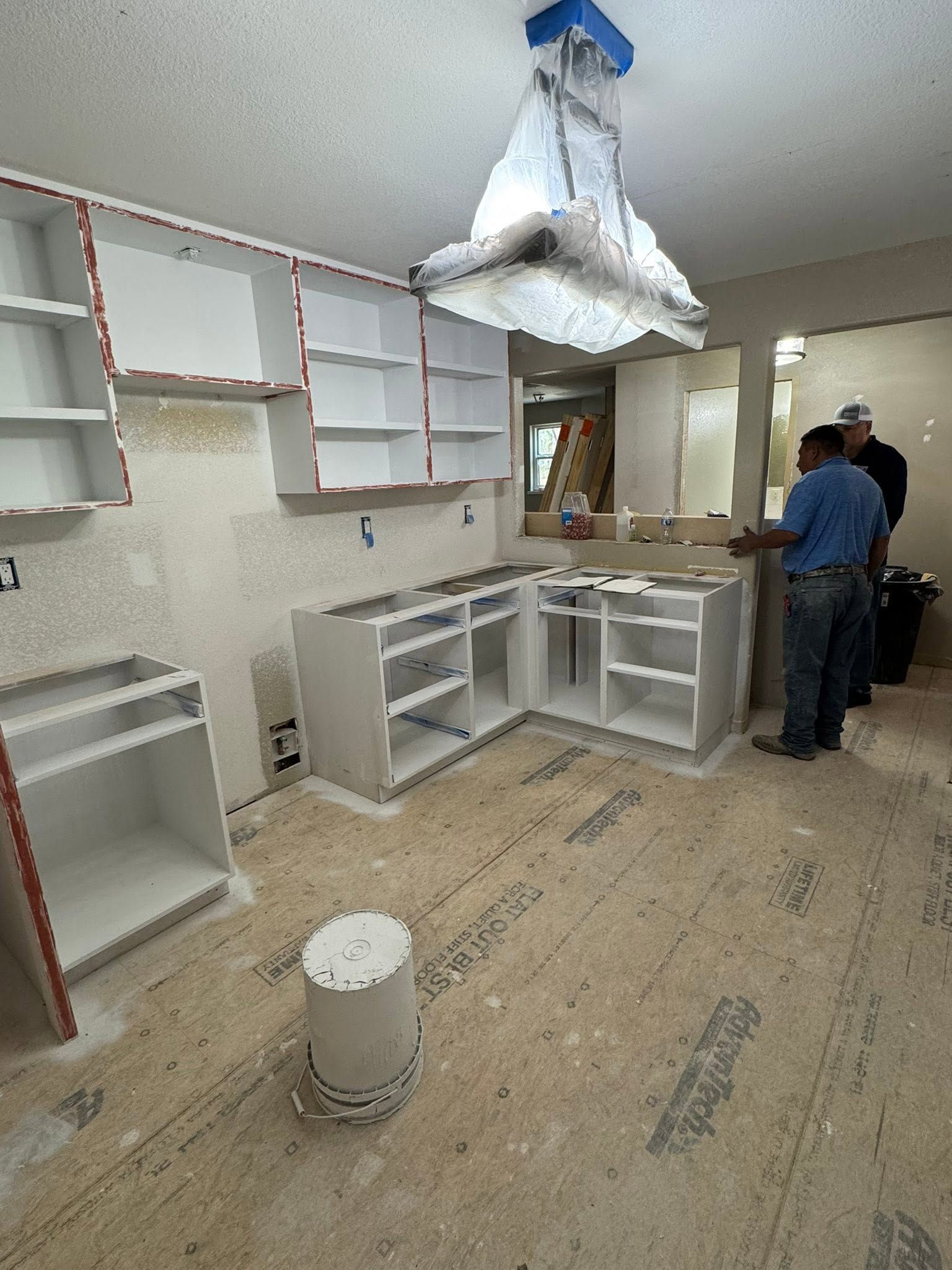 Unfinished kitchen remodel with white cabinets, exposed flooring, and two workers near the sink.