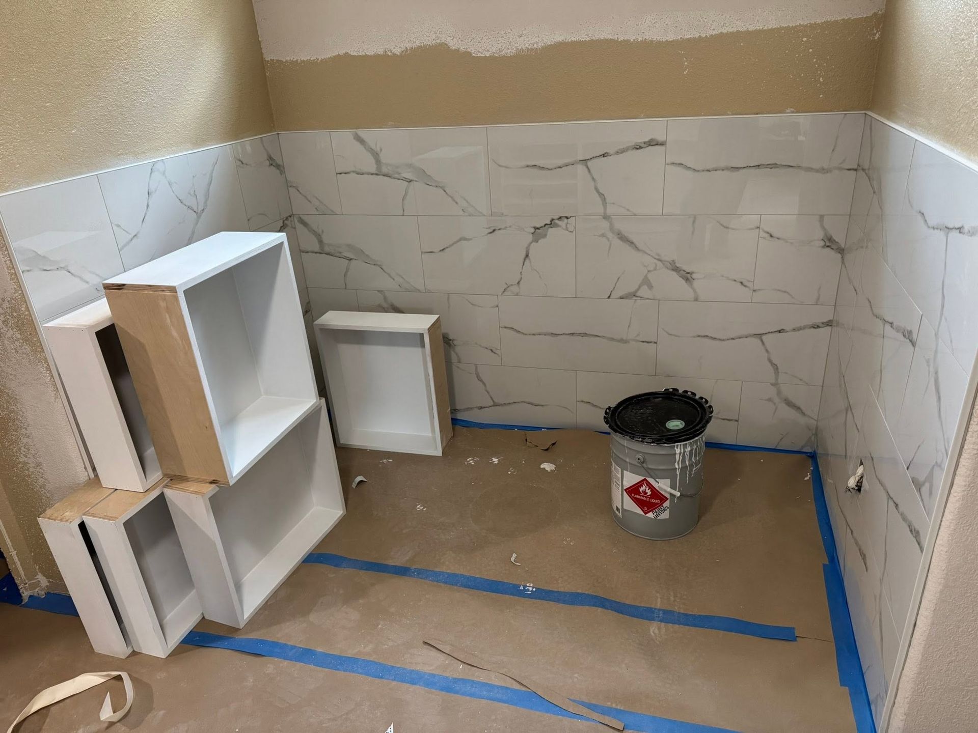 Small tiled room under renovation with white cabinets, blue tape on the floor, and a paint bucket.
