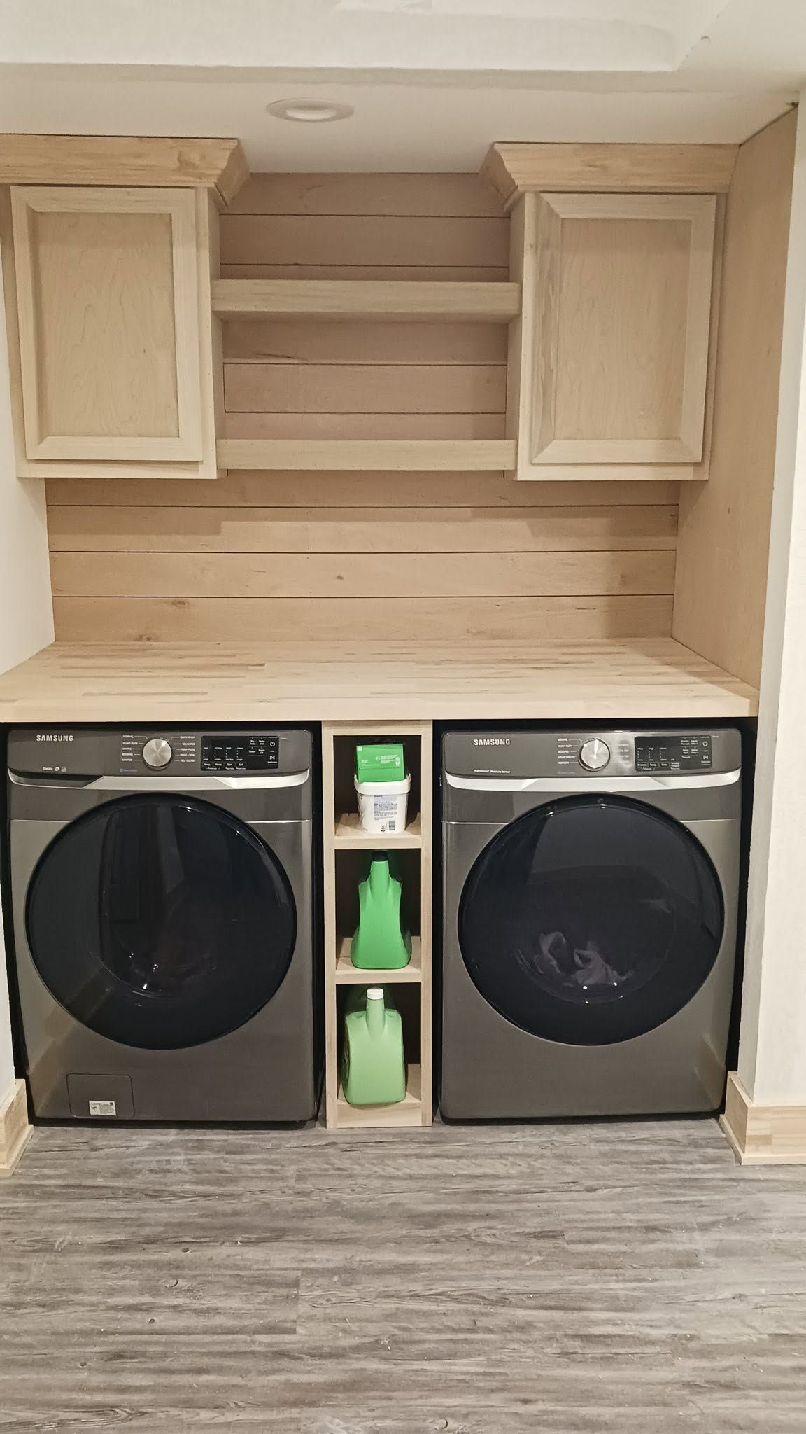 Laundry room with black washer and dryer under beige cabinets and open shelves.