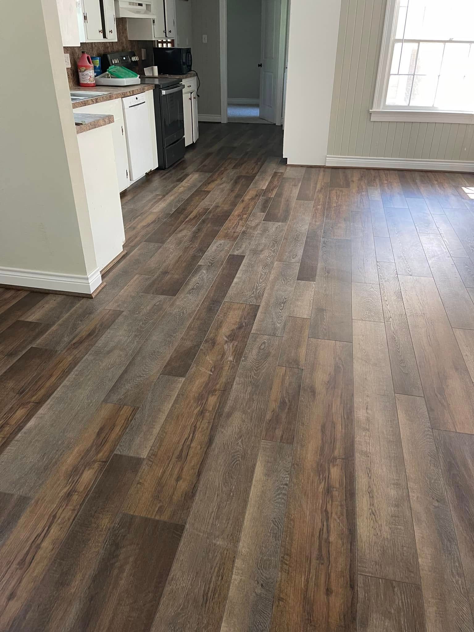 Empty kitchen and living area with wood-look flooring, white cabinets, and a sunlit window.