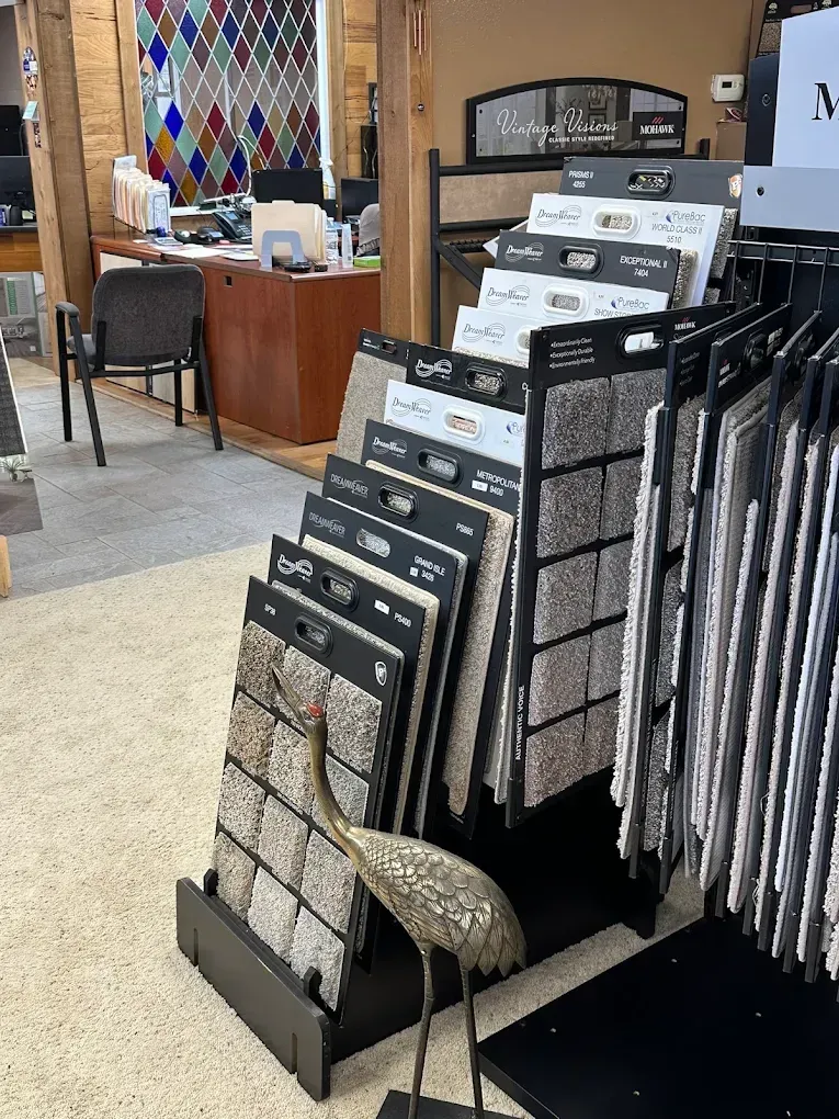 Store display rack with tile samples and carpet swatches in a showroom; a decorative bird figurine sits at the base.