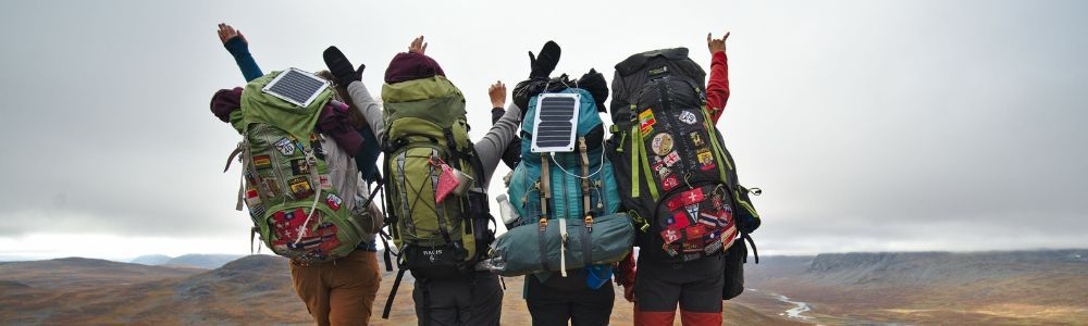 Four hikers with backpacks raise their arms. Each backpack has a solar panel attached. Overcast sky.