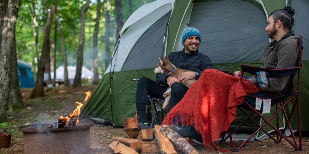 Two men are sitting around a campfire in front of a tent.